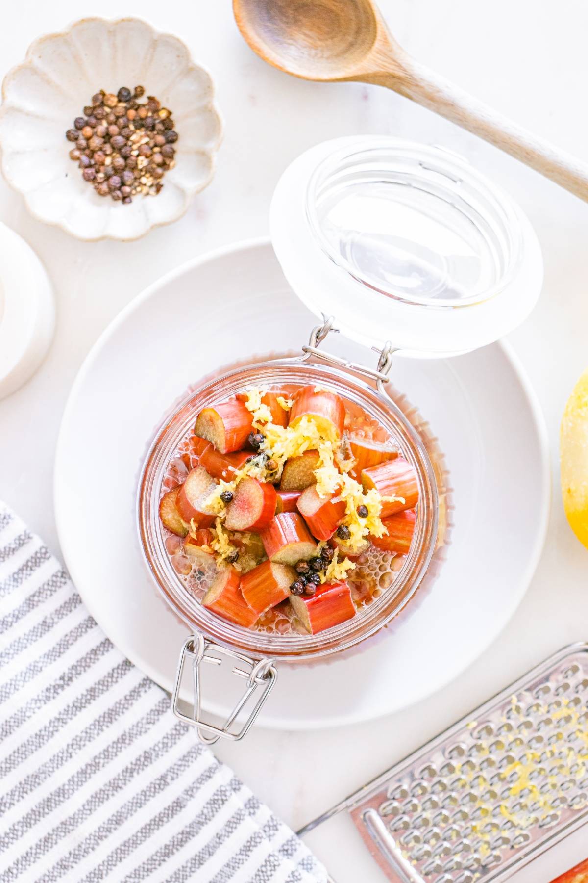 Glass jar with chia pudding topped with chopped rhubarb, lemon zest, and peppercorns on a white plate.