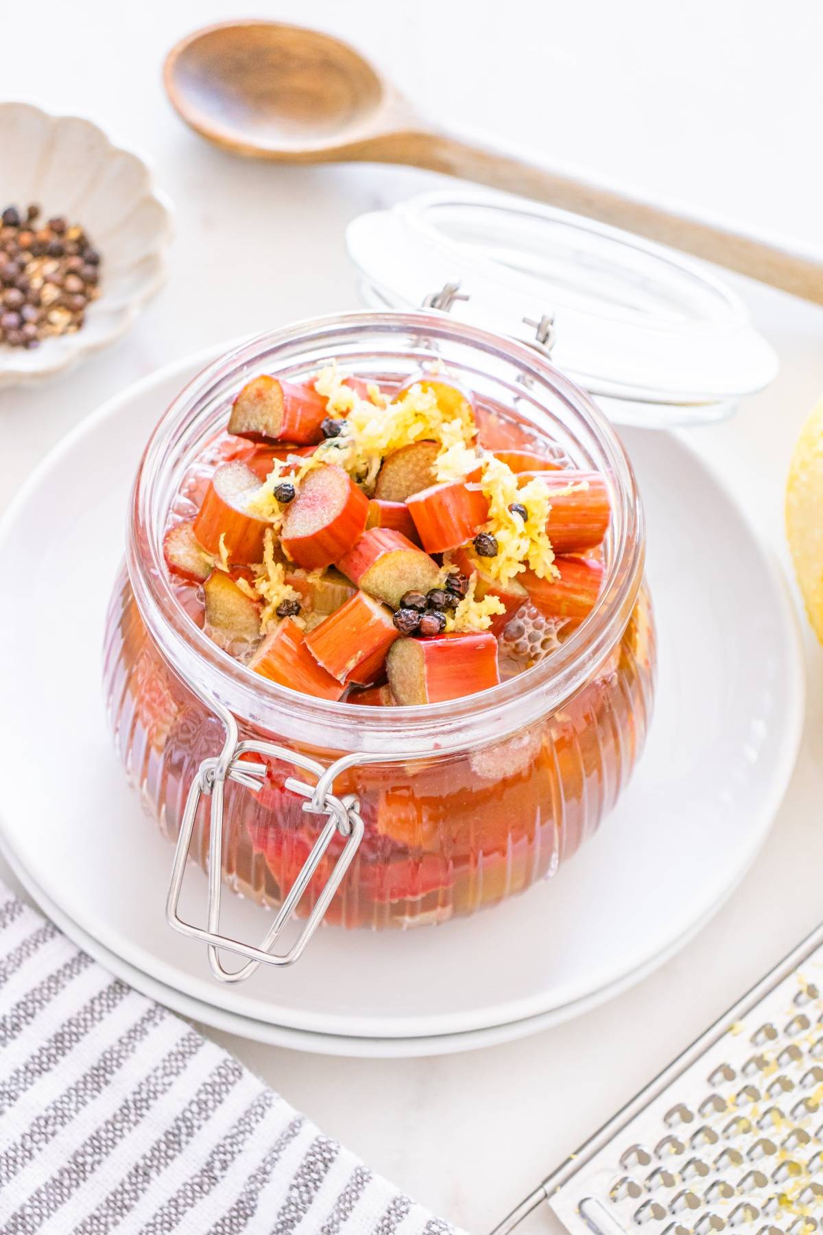 A glass jar filled with chopped rhubarb, lemon zest, and spices sits on a white plate.