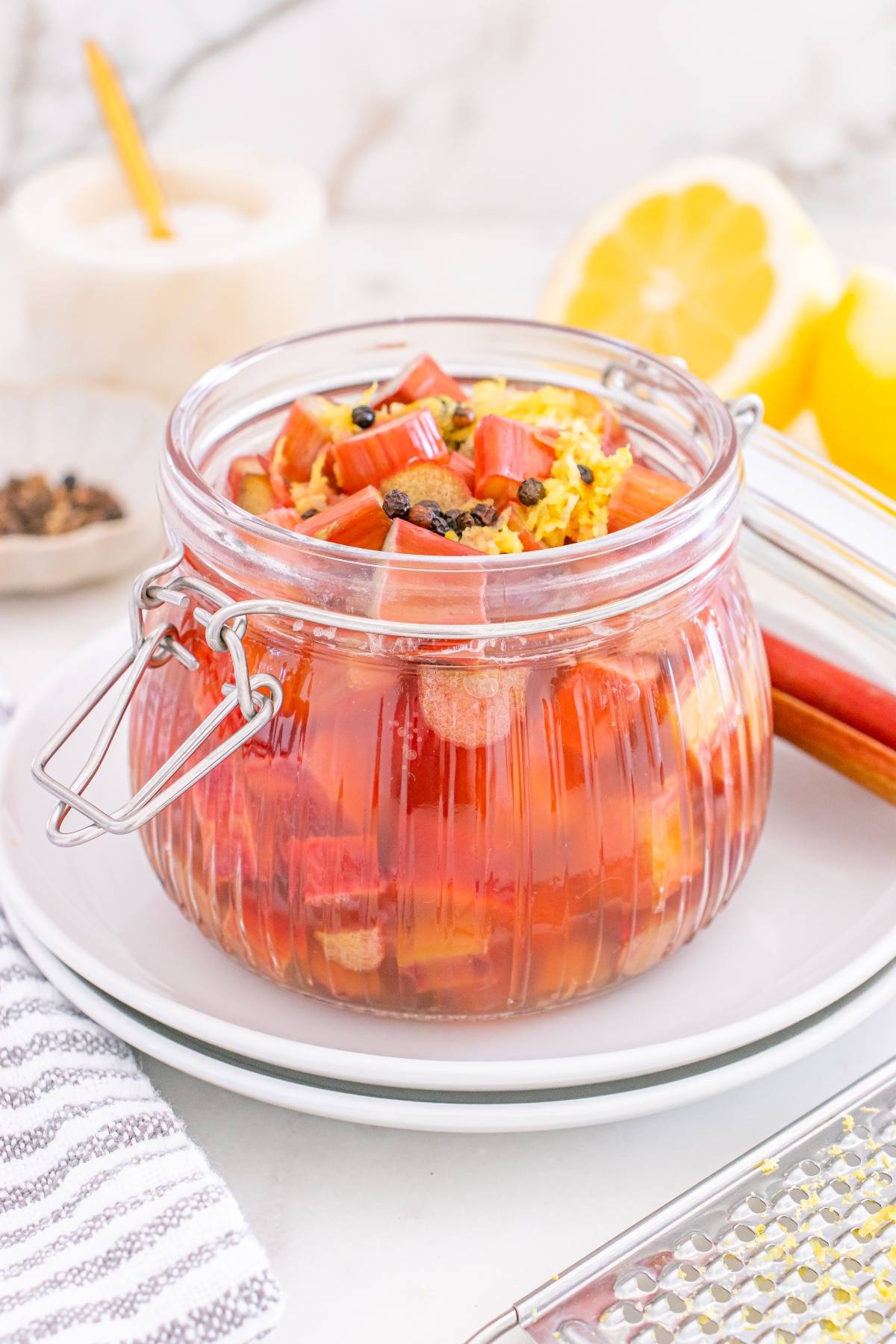 A glass jar filled with chopped rhubarb, lemon zest, and spices sits on a white plate.