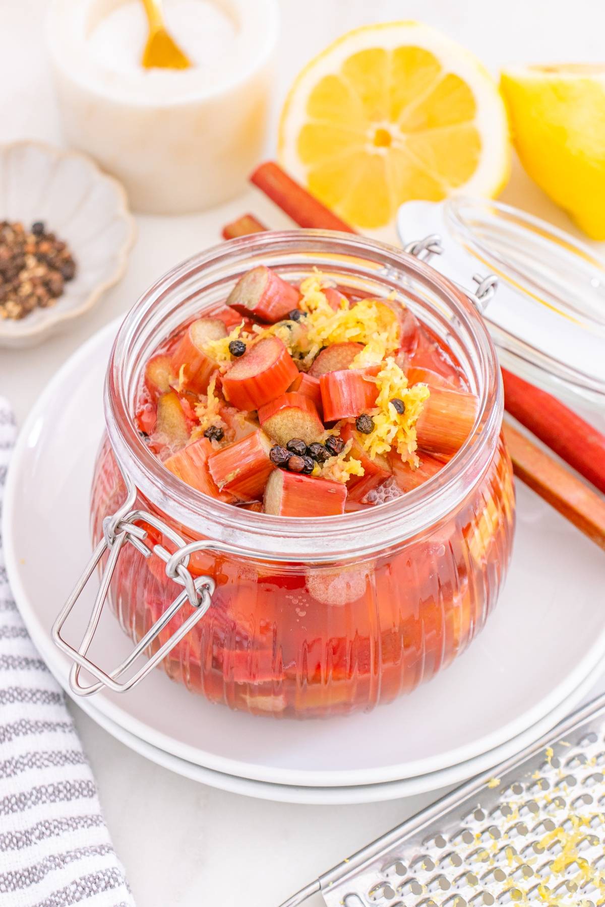 A glass jar of rhubarb compote with lemon zest and spices, surrounded by lemon, cinnamon sticks, and peppercorns.