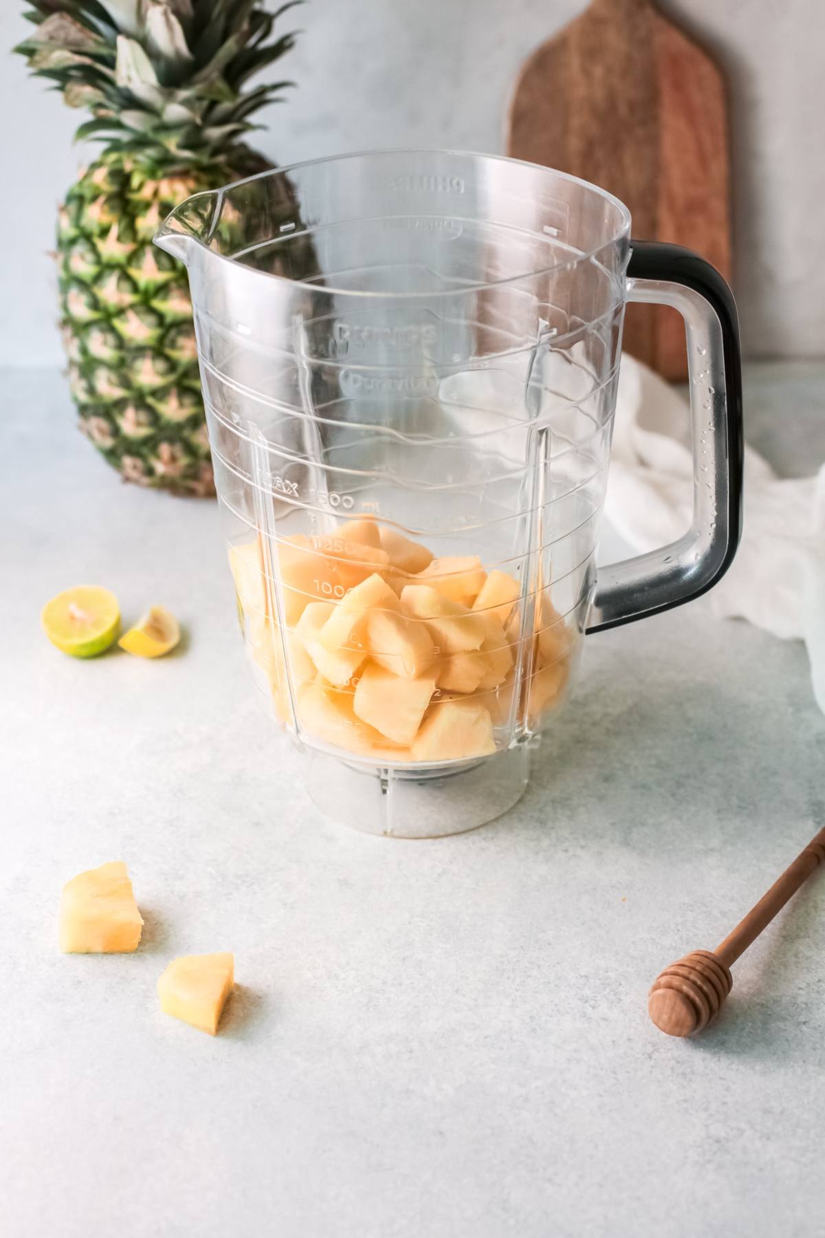 A blender pitcher with pineapple chunks inside, next to a pineapple, lime halves, and honey dipper on a counter—perfect for a refreshing drink recipe or summer drink with pineapple.