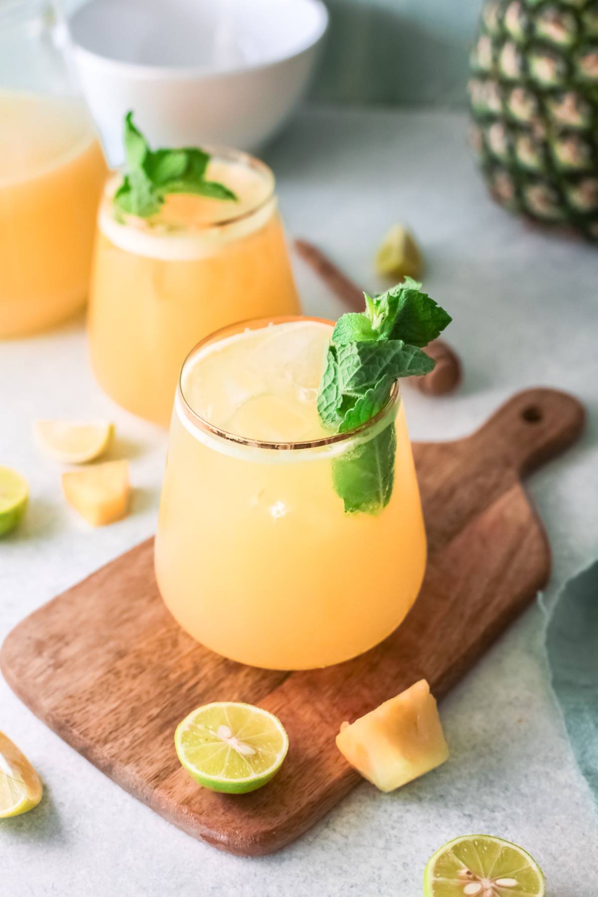A glass of Pineapple Agua Fresca with mint, surrounded by lime halves and pineapple chunks on a wooden board—a refreshing drink recipe perfect for any occasion.