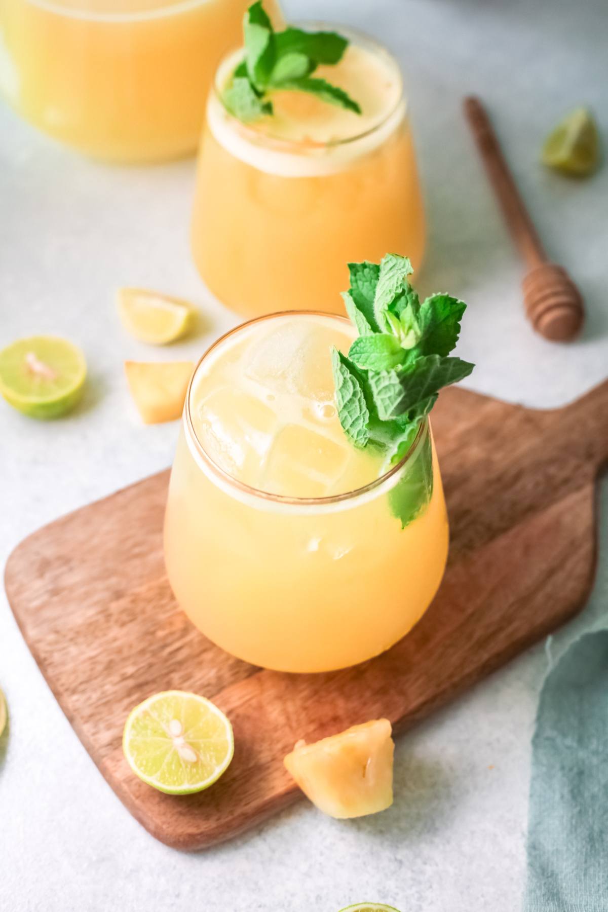 A glass of orange tropical juice with ice and mint on a wooden board, surrounded by lime and pineapple pieces—perfect as a refreshing Summer drink with pineapple or inspired by a Pineapple Agua Fresca recipe.