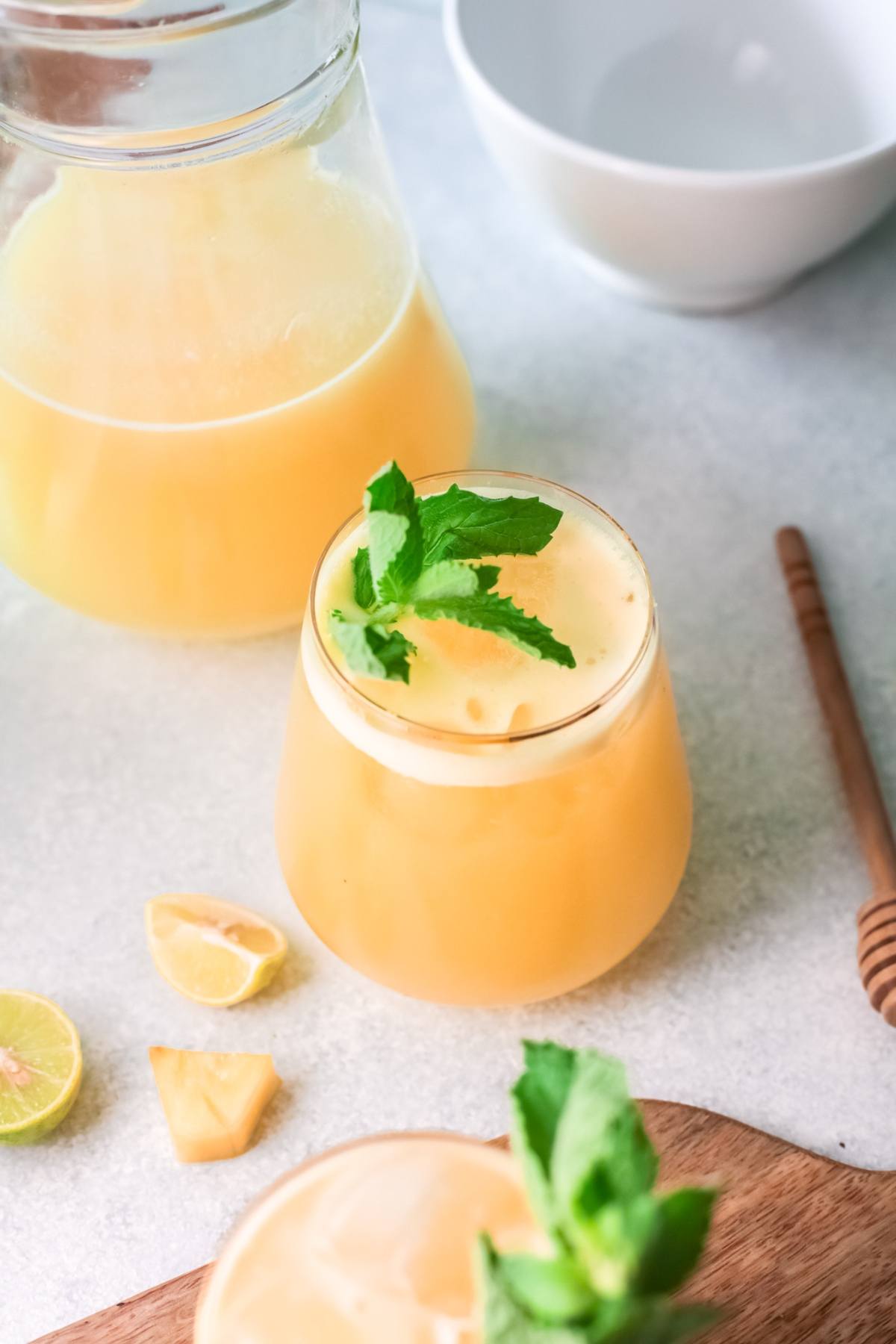 A glass of fresh yellow Pineapple Agua Fresca with mint garnish sits by a jug, lime wedges, and a honey dipper—a refreshing non-alcoholic drink recipe perfect for summer.