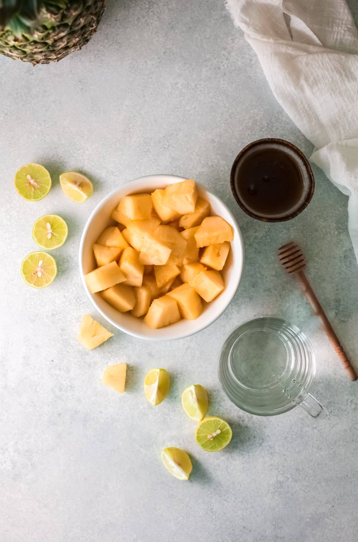 Bowl of pineapple chunks with lime halves, honey dipper, glass pitcher, and a small bowl of honey—perfect ingredients for making a refreshing drink recipe like Pineapple Agua Fresca on a light surface.