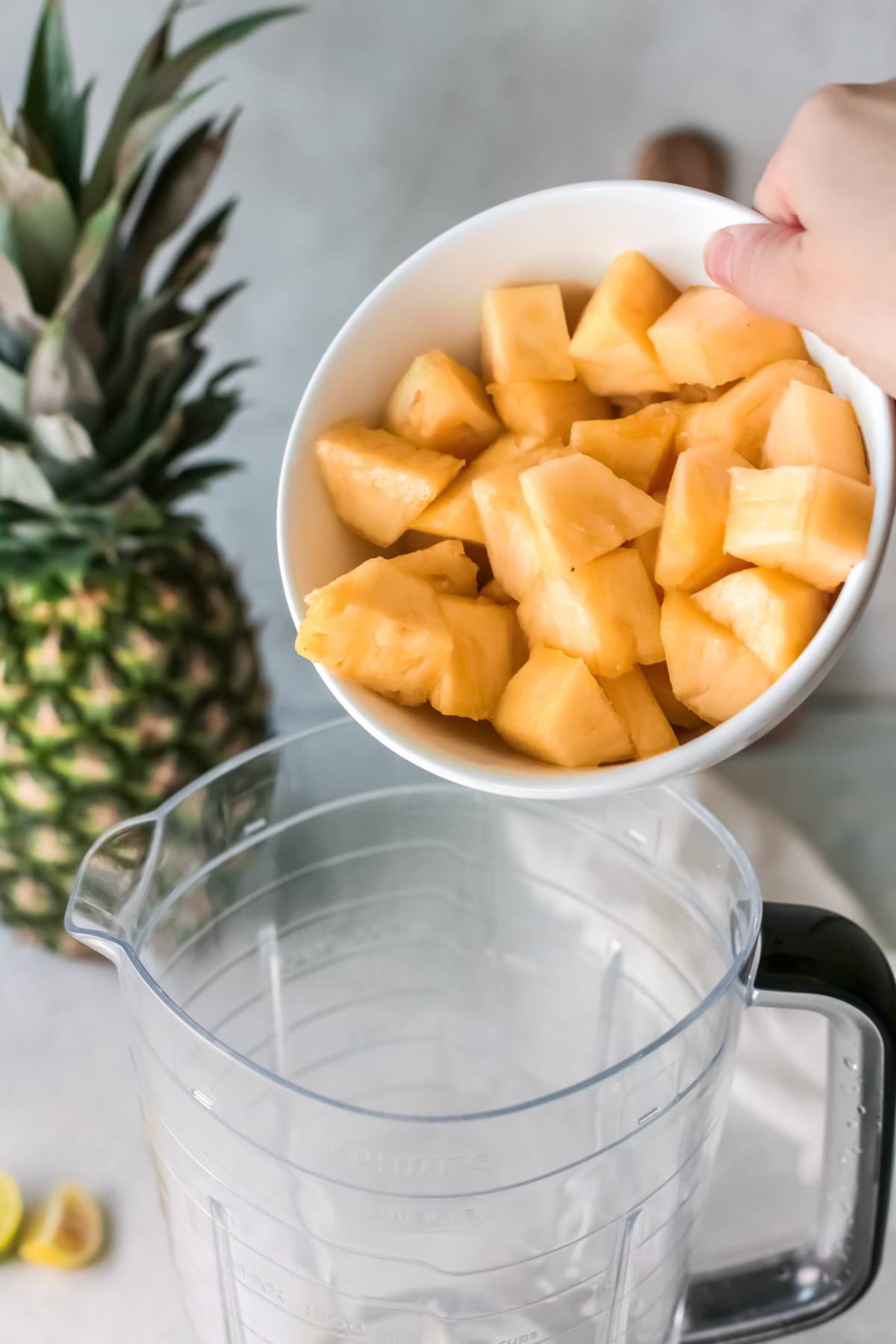A hand pours pineapple chunks from a bowl into a blender, preparing a refreshing drink recipe with a whole pineapple nearby.