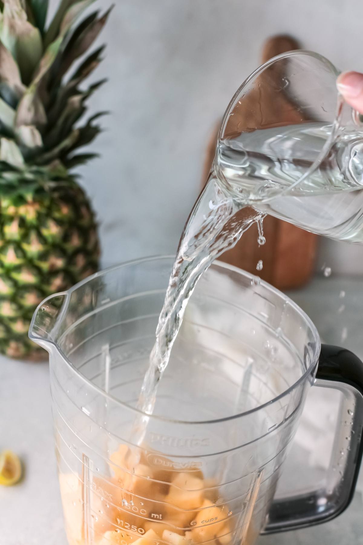 A hand pours water from a glass into a blender with pineapple chunks, next to a whole pineapple—creating a refreshing drink recipe perfect for Pineapple Agua Fresca.