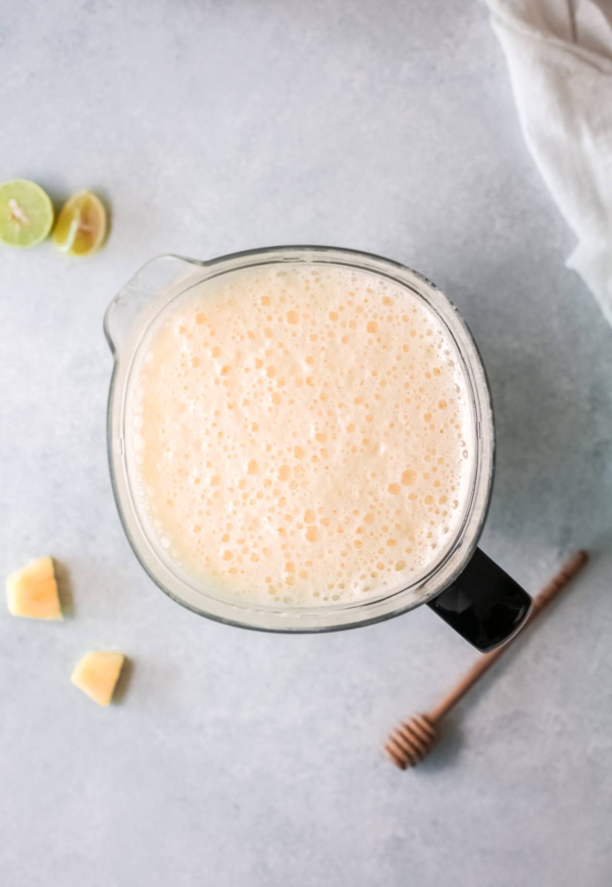 A blender filled with a frothy smoothie, surrounded by lime, pineapple pieces, and a honey dipper—perfect inspiration for your next refreshing drink recipe.