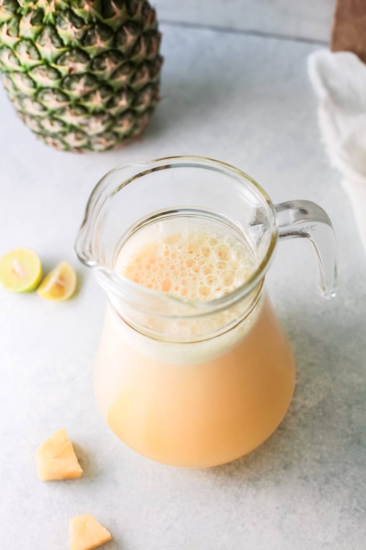 A glass pitcher of pineapple juice on a light surface with pineapple, lime, and pineapple pieces nearby—a perfect summer drink with pineapple for a refreshing treat.