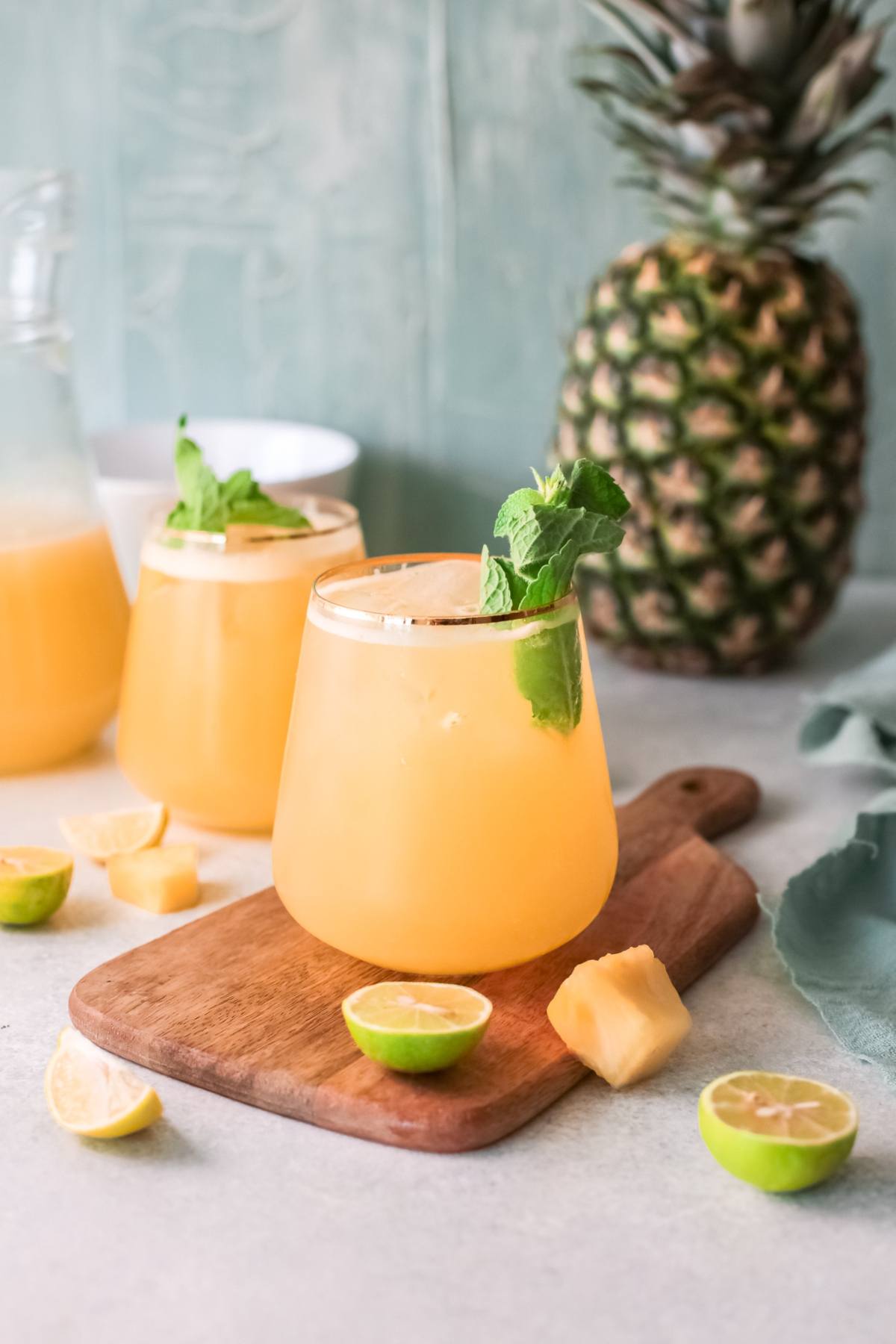 Two glasses of summer drink with pineapple, lime, and mint sit on a wooden board, with a whole pineapple in the background—a perfect refreshing drink recipe for warm days.