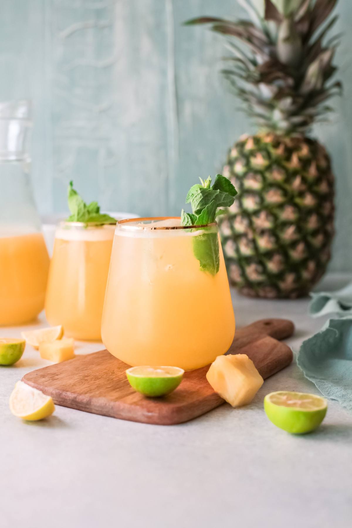 Two glasses of a summer drink with pineapple, mint, lime wedges, and pineapple pieces on a wooden board beside a whole pineapple—a refreshing drink recipe perfect for warm days.