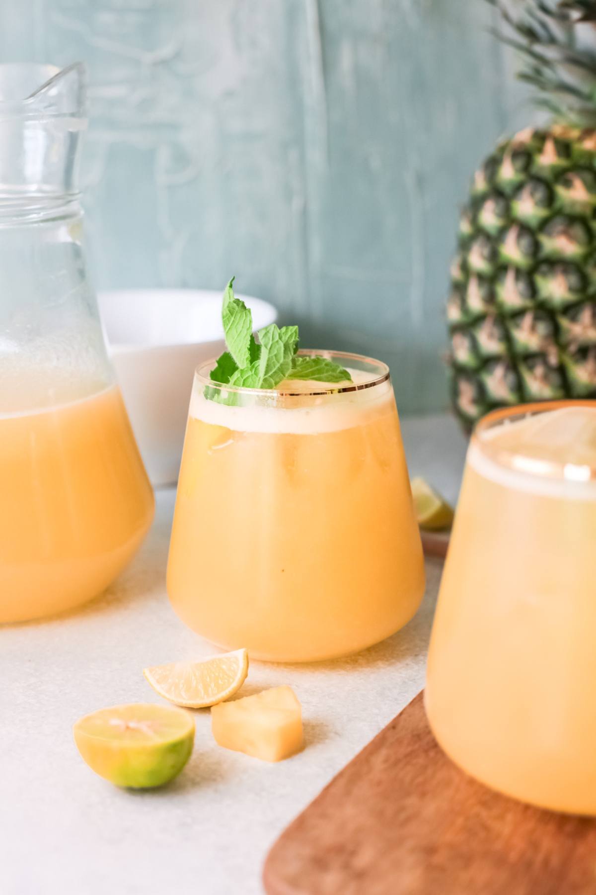 A glass of pineapple juice with mint, surrounded by pineapple, lime wedges, and a jug of juice—a perfect summer drink with pineapple for a refreshing non-alcoholic drink recipe.