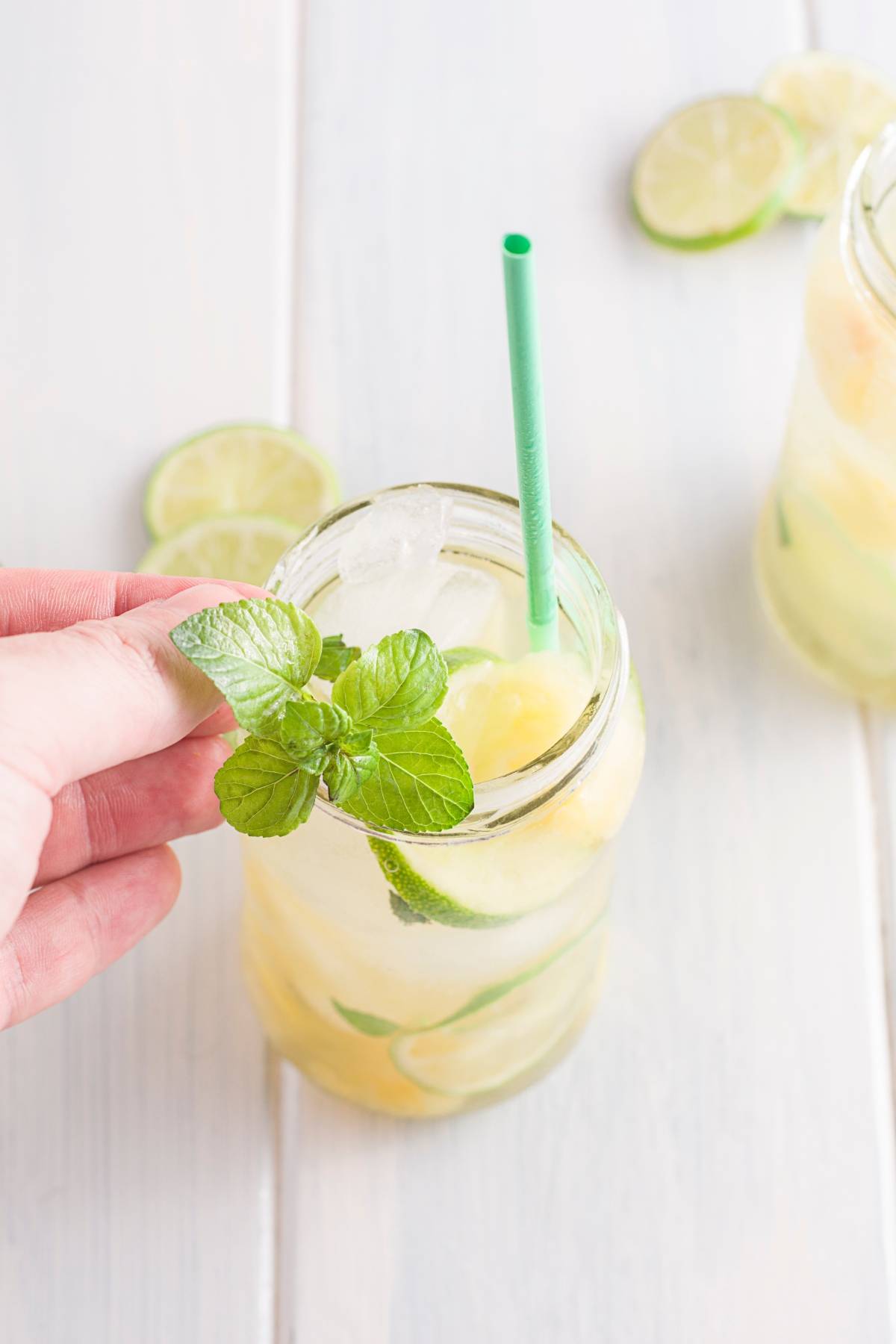 A hand holding a glass of lemonade with ice, lime slices, mint, and a green straw on a light surface—perfect inspiration for your next pineapple ginger mojito mocktail.