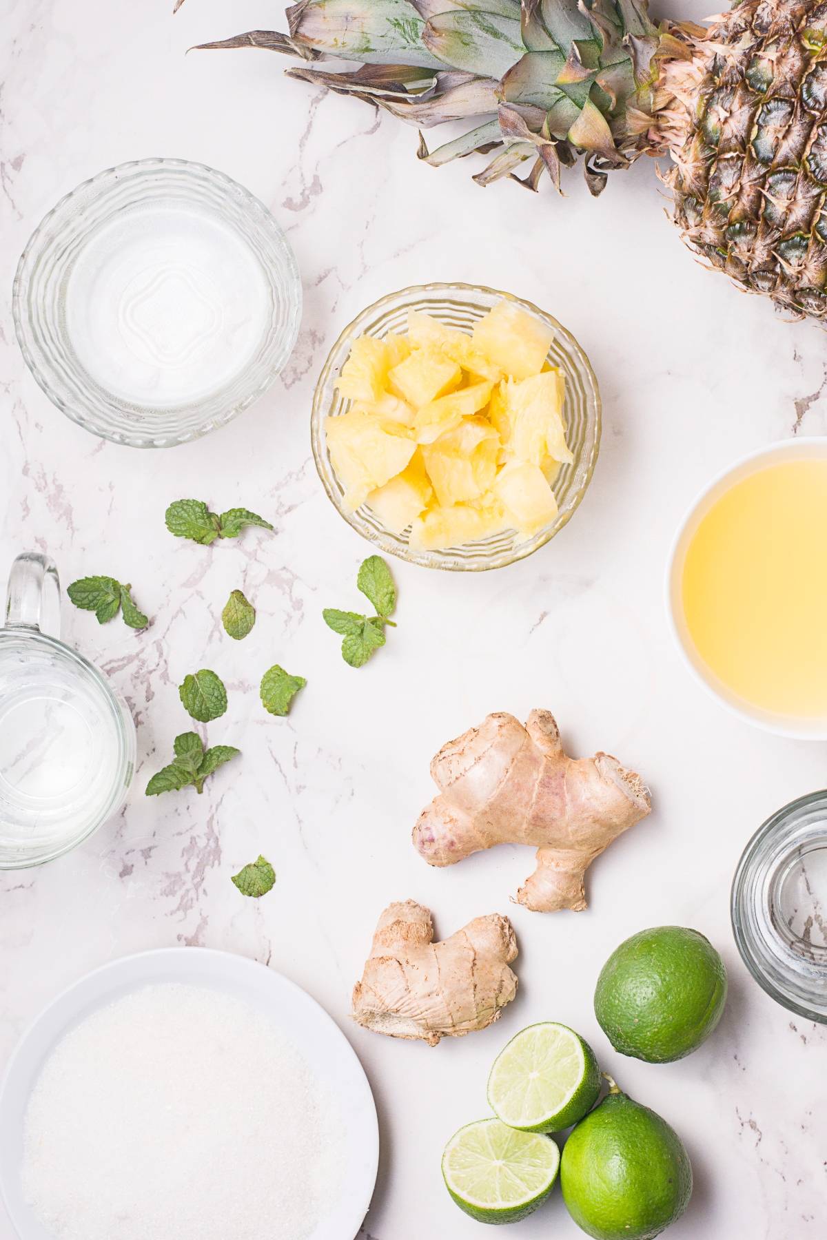 Sliced pineapple, limes, ginger, mint, and sugar sit beside glasses of juice and water on a white marble surface—perfect for trying an easy pineapple mojito or a refreshing pineapple ginger mocktail.