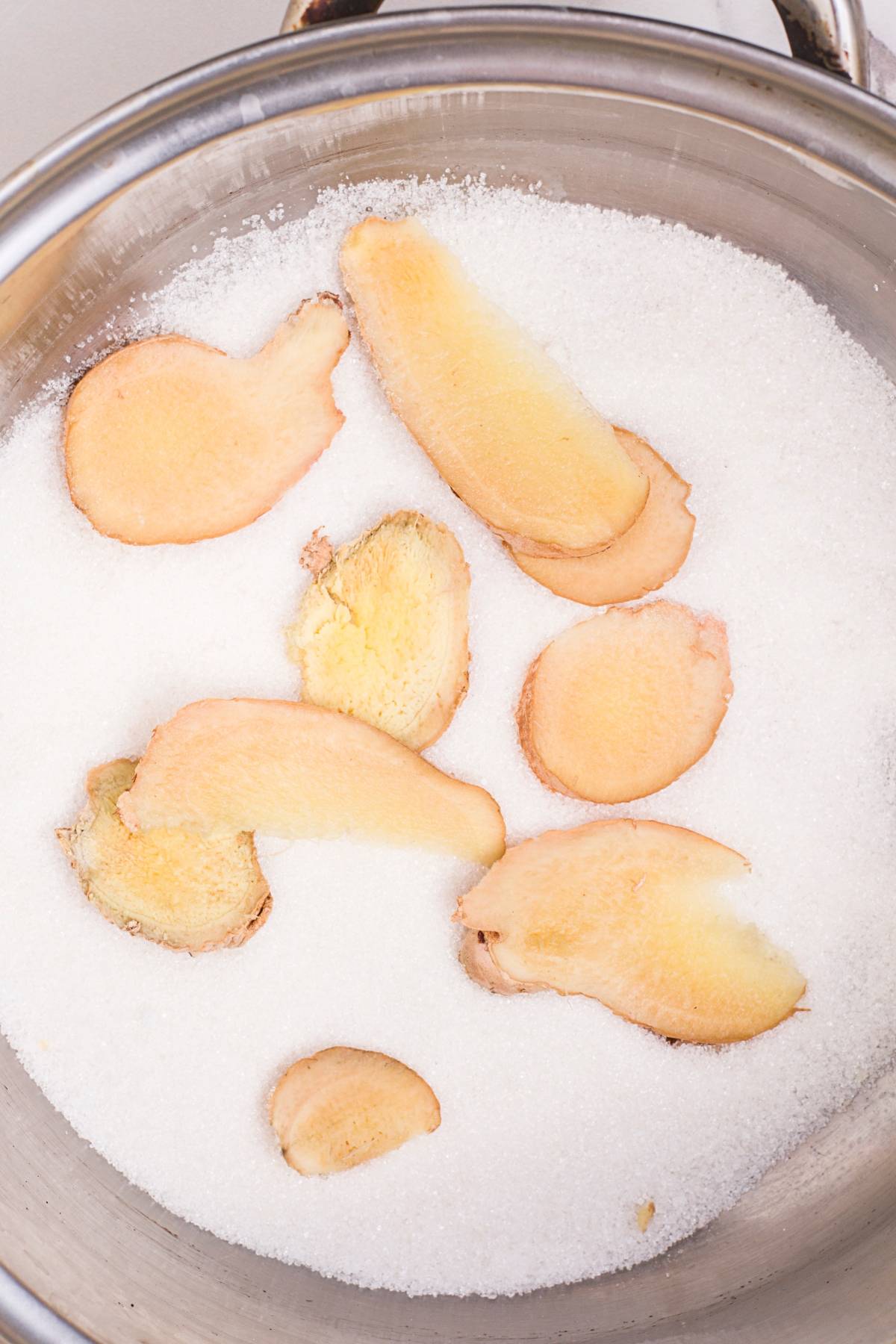 Sliced fresh ginger on a bed of granulated sugar in a metal bowl, perfect for infusing your Pineapple ginger mocktail with vibrant flavor.