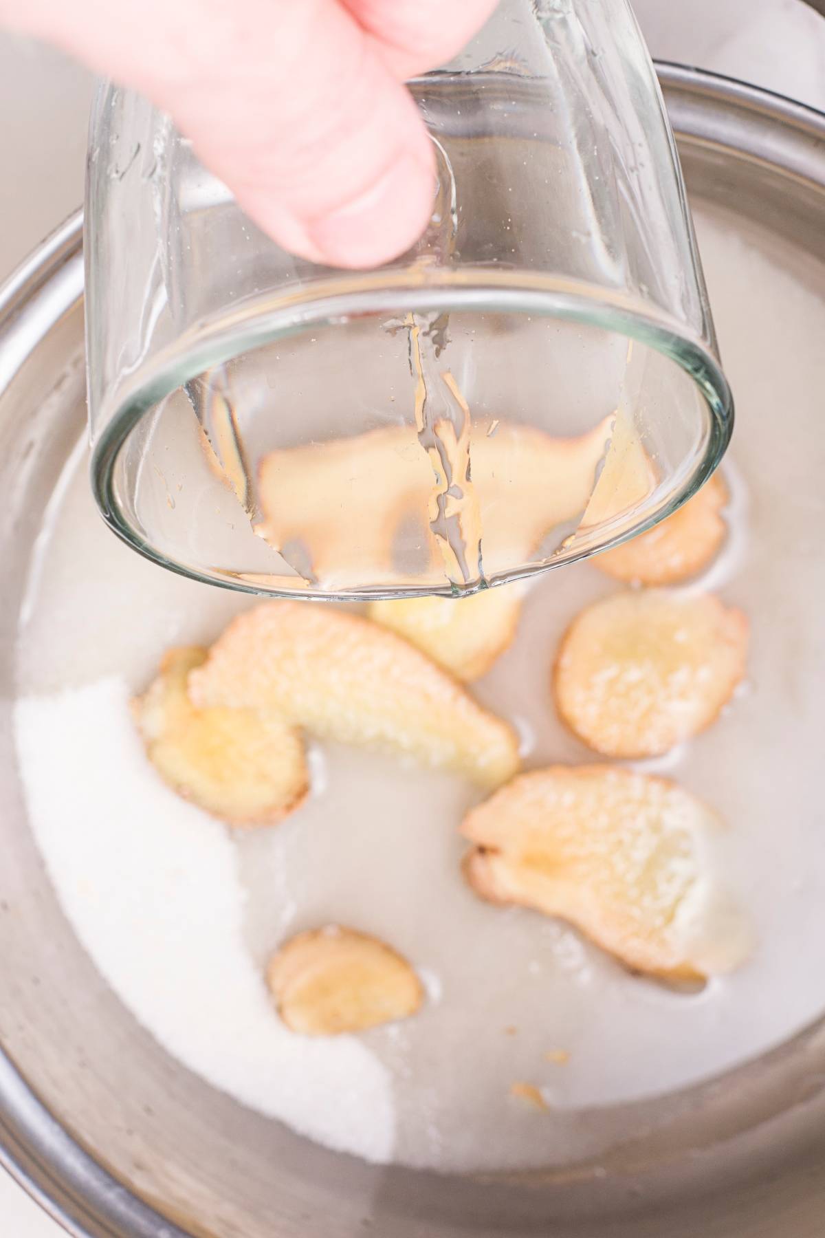 A hand pours liquid from a glass into a bowl with ginger slices and sugar—perfect for preparing an easy pineapple mojito or a refreshing pineapple ginger mocktail.