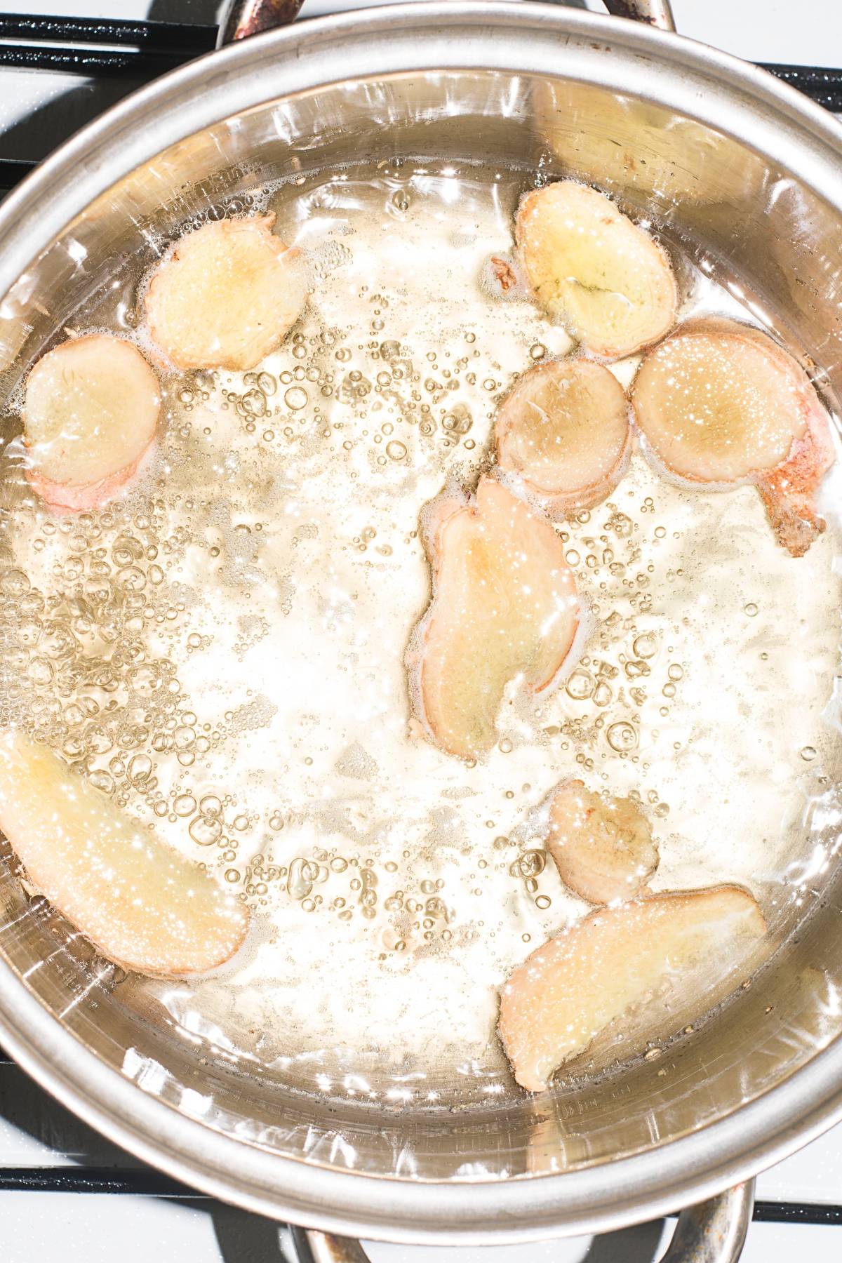 Sliced ginger frying in bubbling oil inside a stainless steel pan on a stove, infusing the air with an aroma perfect for a refreshing Pineapple ginger mojito mocktail.