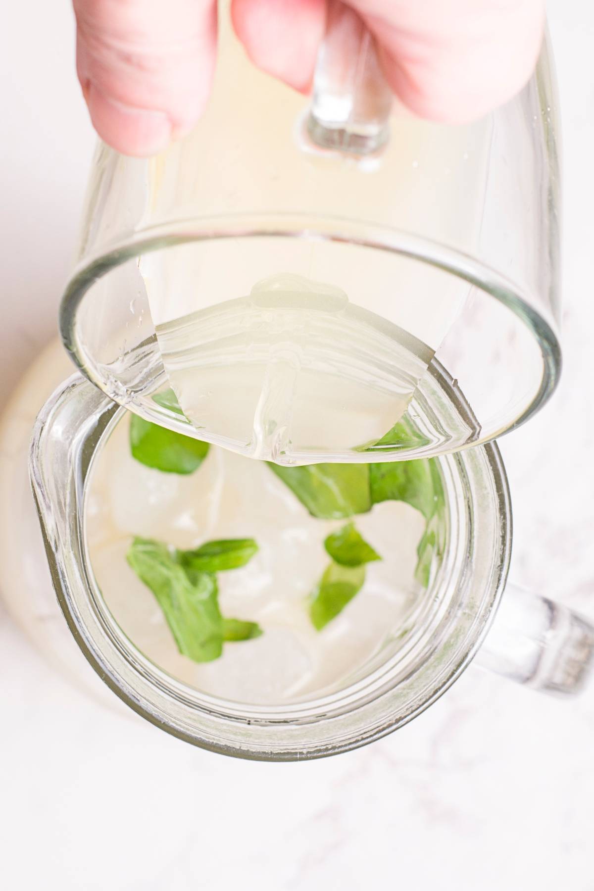 A hand pours a light-colored tropical mocktail with ice and mint leaves into a glass mug from above.