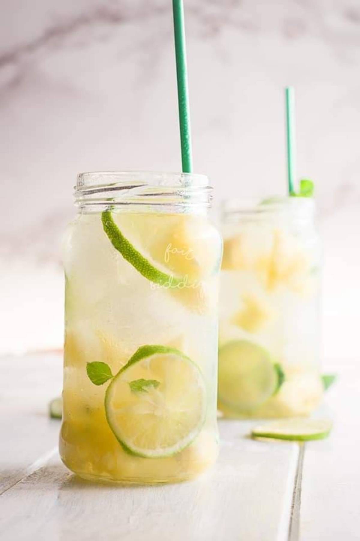 Two mason jar glasses of a refreshing Pineapple ginger mocktail with lime slices, ice, and green straws sit on a light wooden surface.