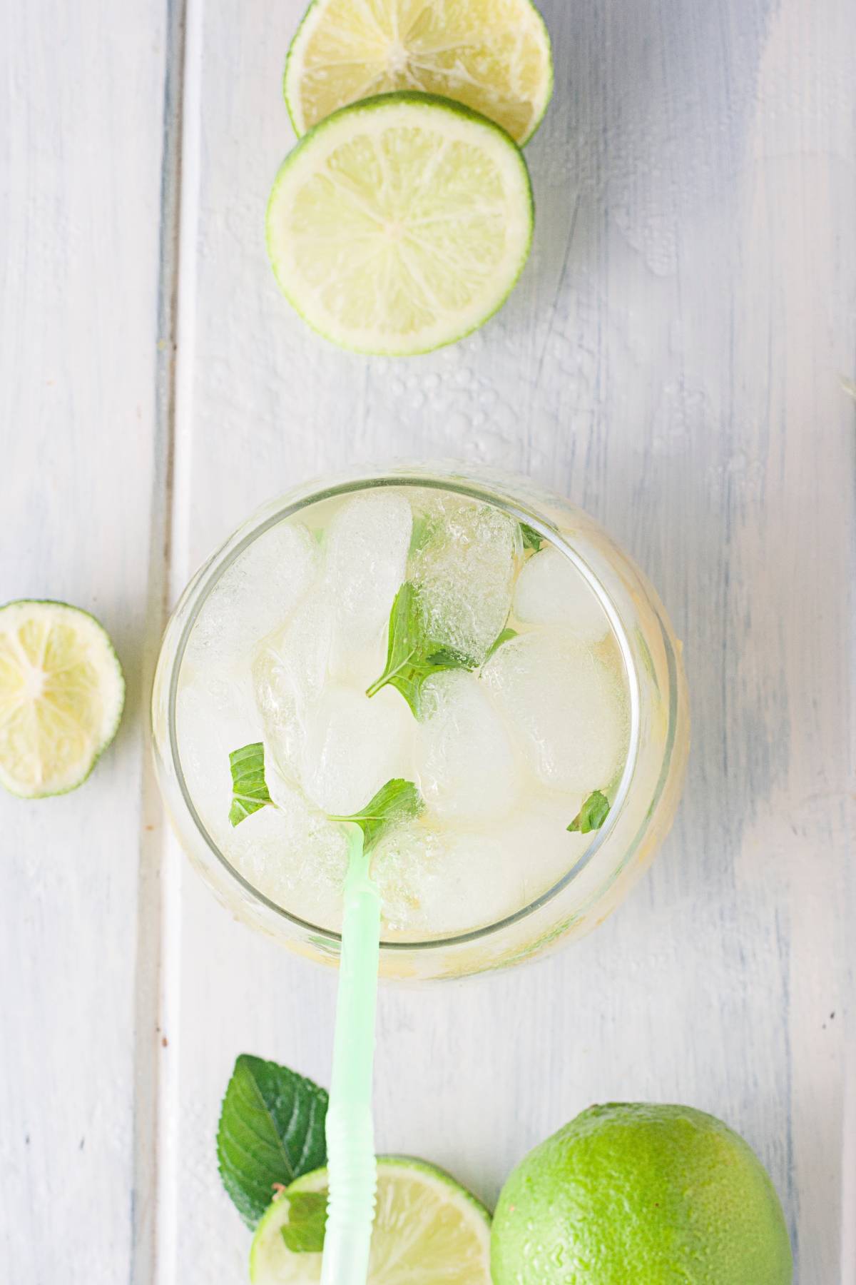 Top view of a Pineapple ginger mojito mocktail with ice, mint leaves, and a green straw, surrounded by fresh lime slices on a white surface—a refreshing tropical mocktail experience.