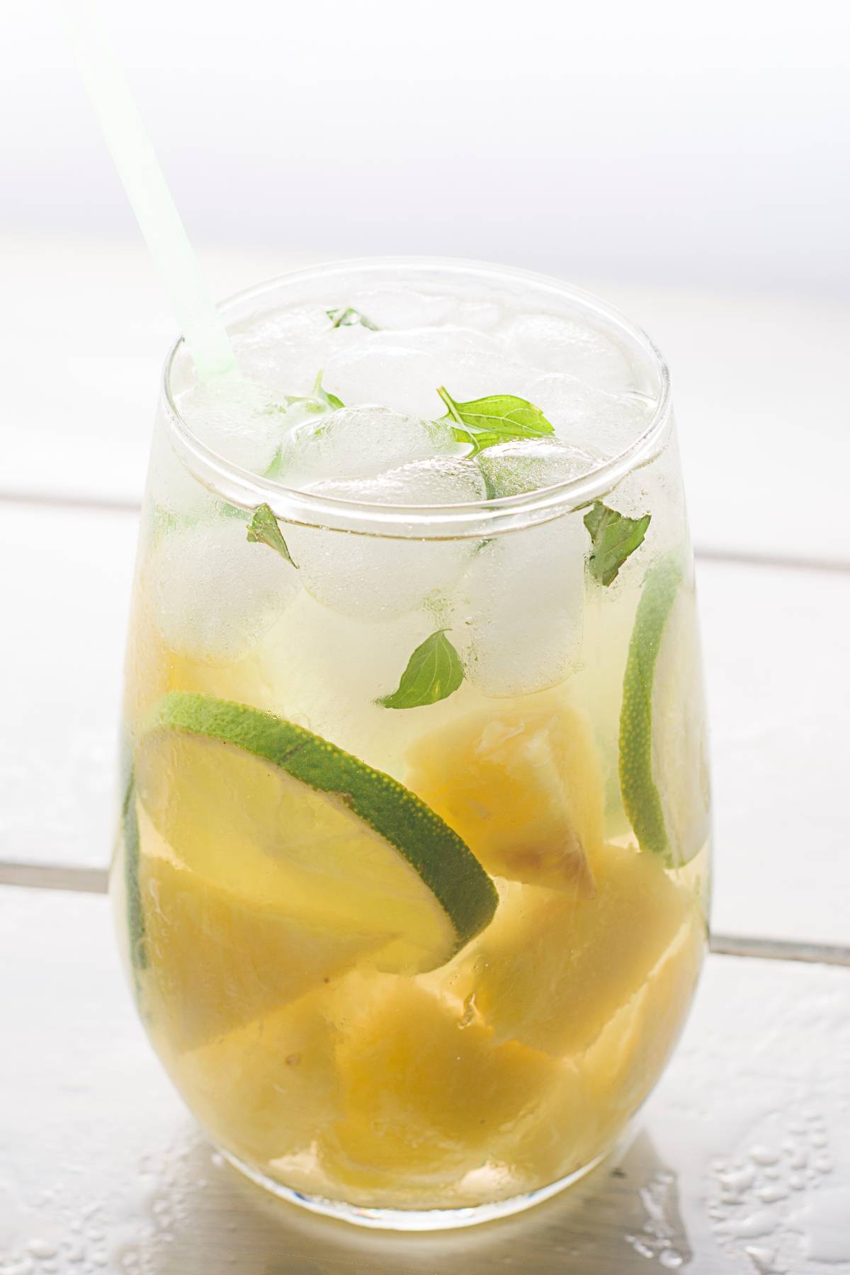 Glass of iced Pineapple ginger mojito mocktail with lime slices, pineapple chunks, mint leaves, and a straw on a light surface.
