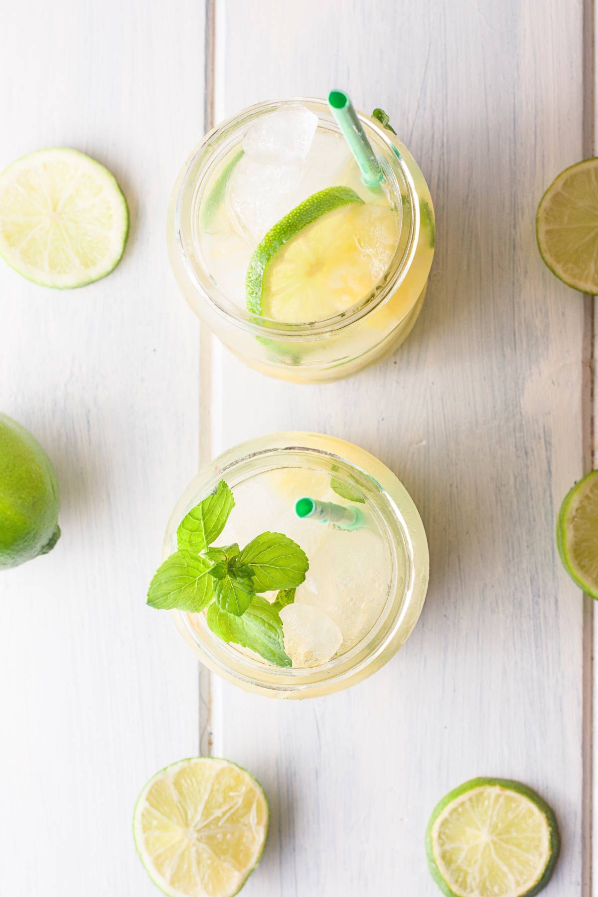Two glasses of iced limeade with green straws, lime slices, and mint, surrounded by lime slices on a white table—perfect inspiration for your next tropical mocktail or easy pineapple mojito.