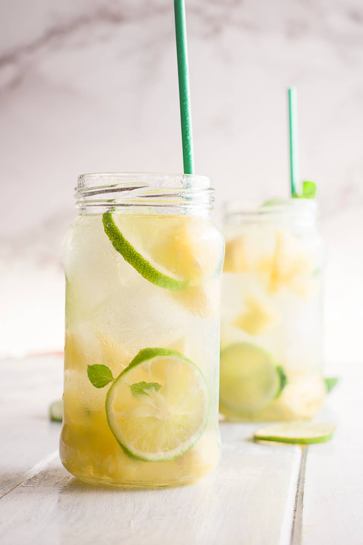 Mason jars of ice water with lime slices, mint, and green straws on a light wooden surface—perfect inspiration for your next Pineapple ginger mojito mocktail.