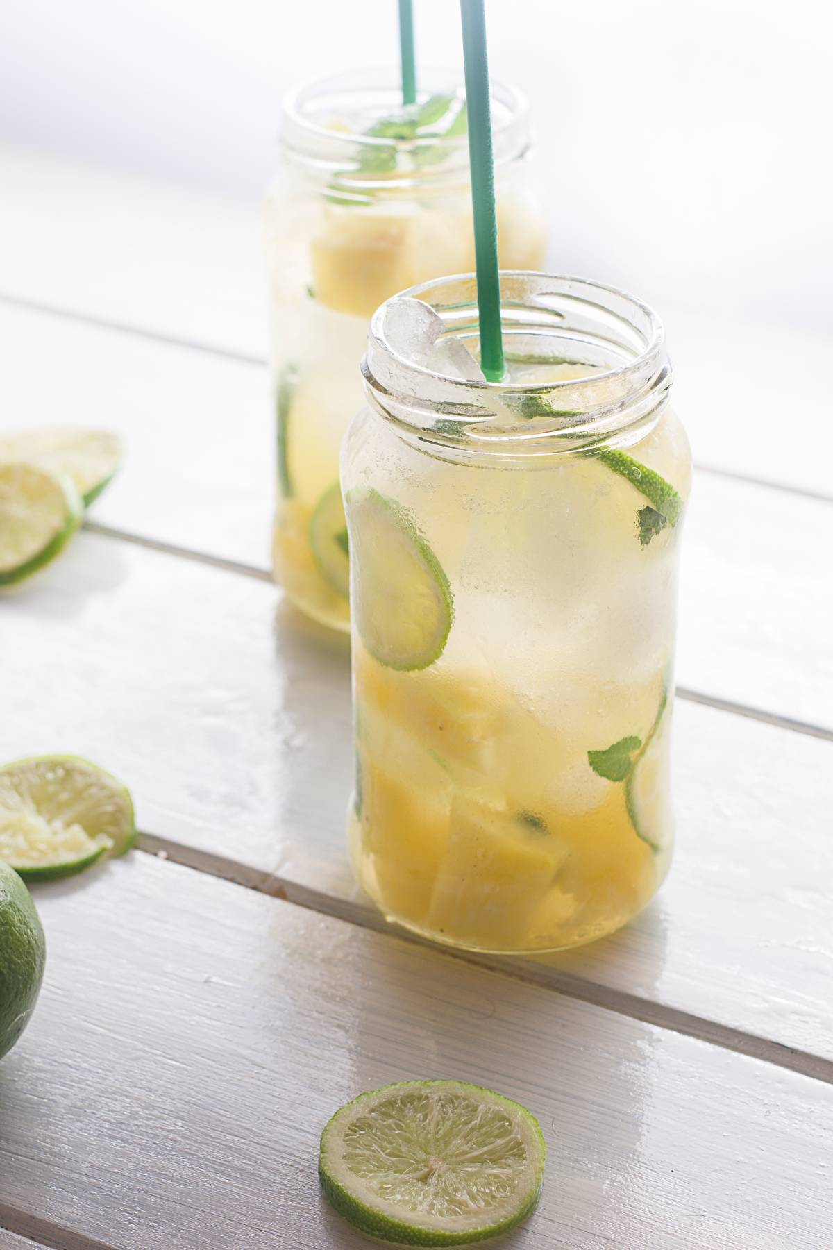 Two mason jars with refreshing Pineapple ginger mojito mocktail, garnished with lime slices and straws, sit on a white wooden surface.