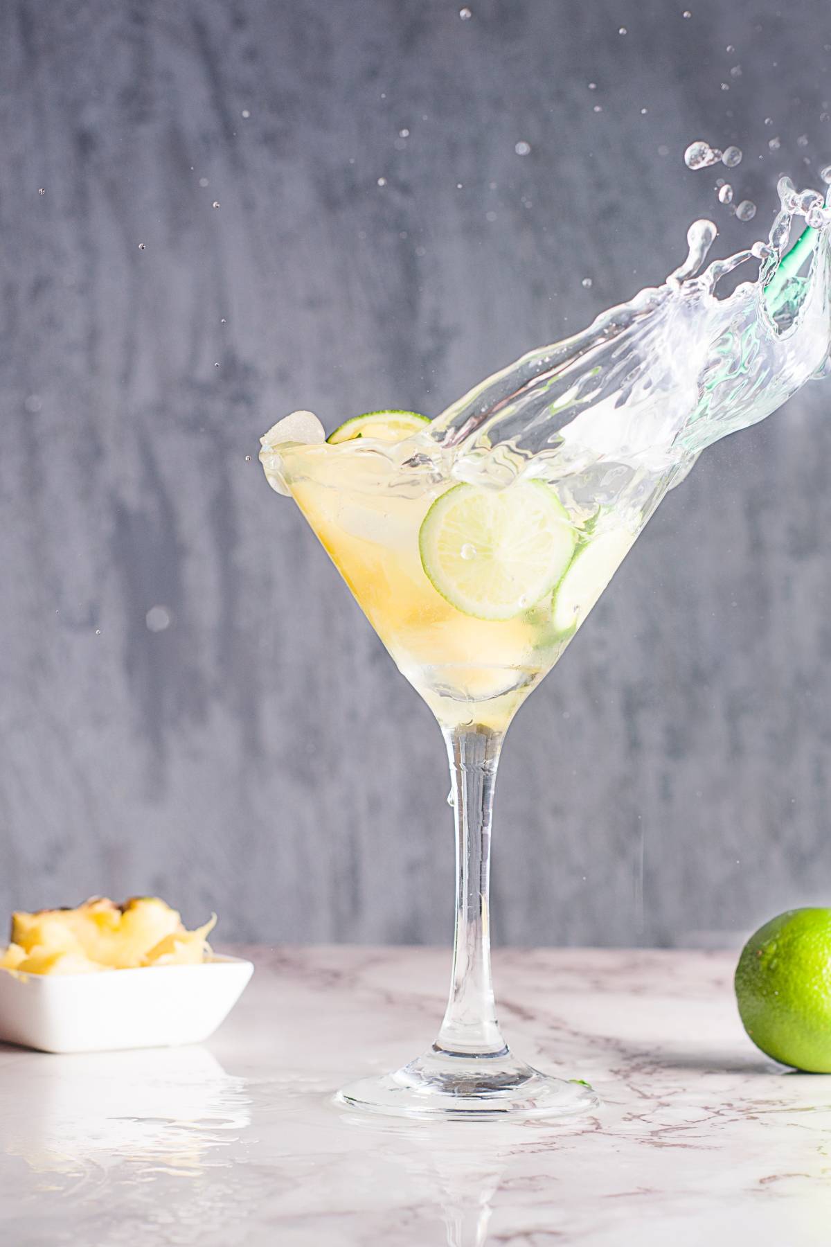 A martini glass with lime slices splashes as liquid spills out, surrounded by lime and pineapple pieces—perfect for serving a refreshing Pineapple ginger mocktail or an easy pineapple mojito.