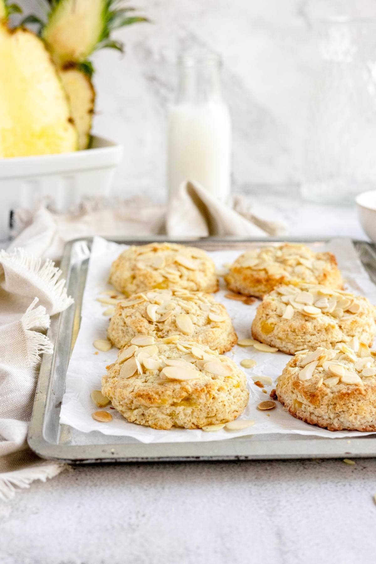 Six almond-topped scones on a baking tray, with a bottle of milk and pineapple in the background—perfect for those seeking scones made with pineapple or looking for tips for making scones at home.