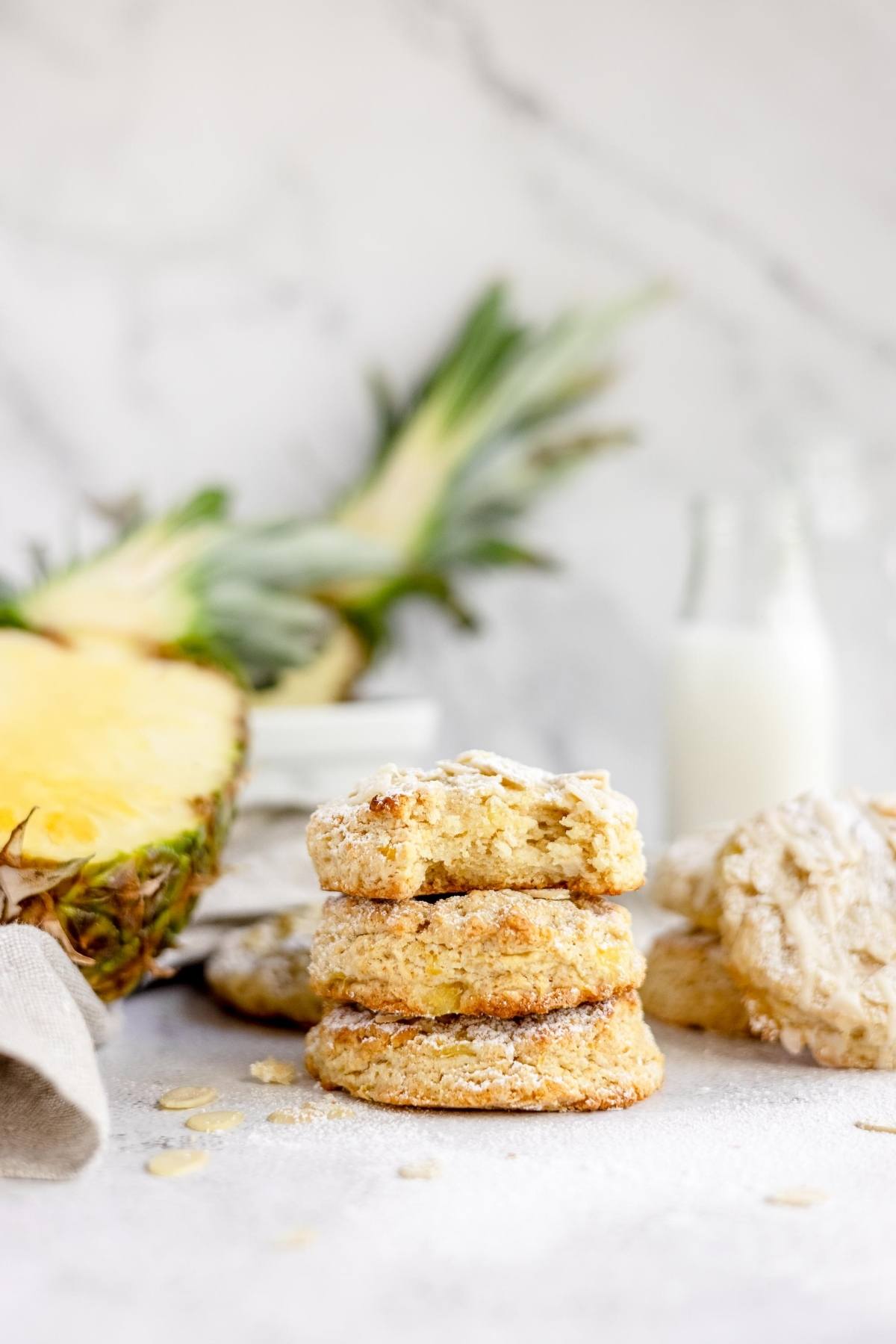 Three scones stacked in front of a sliced pineapple, with a milk bottle in the background on a white surface—perfect inspiration for your next recipe using crushed pineapple or to try the best fruit scone recipe.