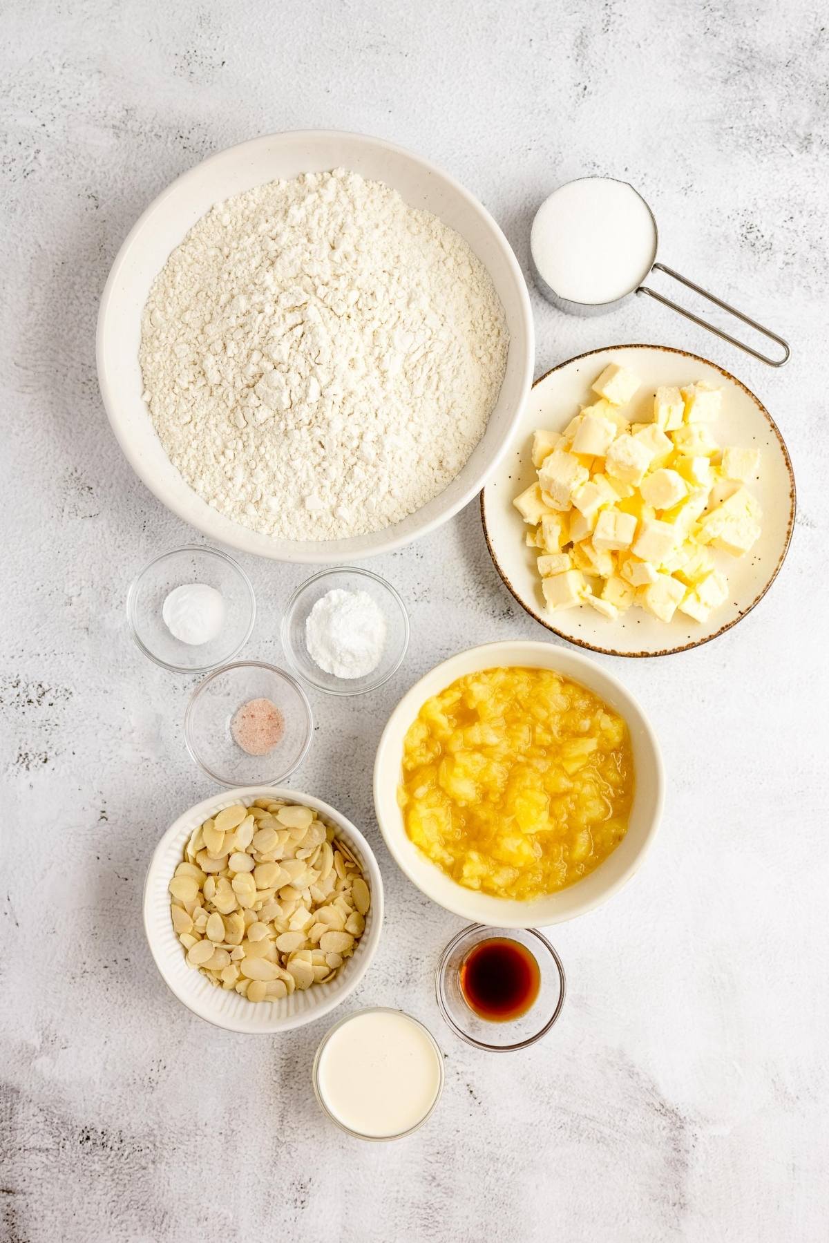 Baking ingredients on a white surface: flour, sugar, butter, sliced almonds, vanilla, milk, salt, and crushed pineapple—perfect for the best fruit scone recipe or making delicious pineapple scones.