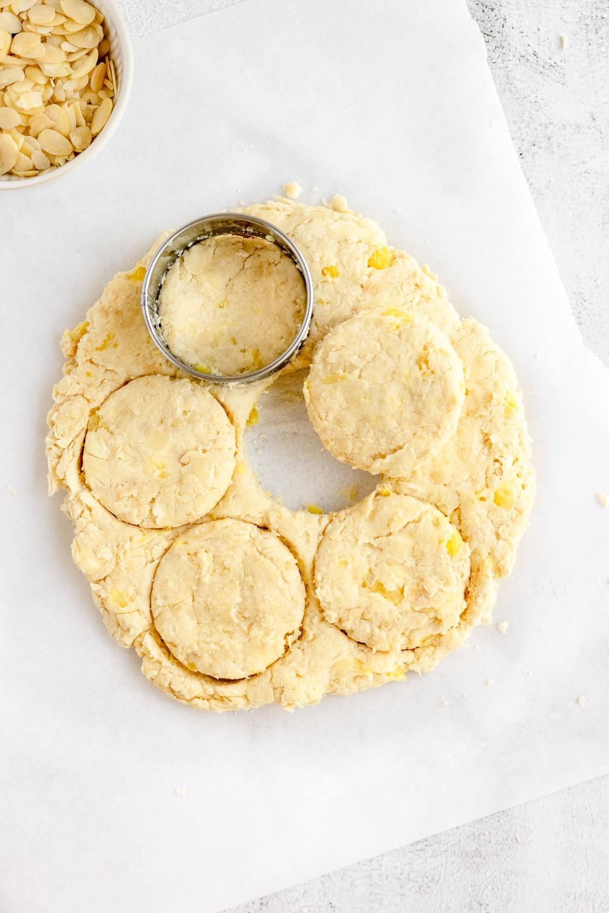 Biscuit dough being cut into rounds with a metal cutter on parchment paper, bowl of almond slices nearby—perfect for scones made with pineapple or for trying a new recipe using crushed pineapple.