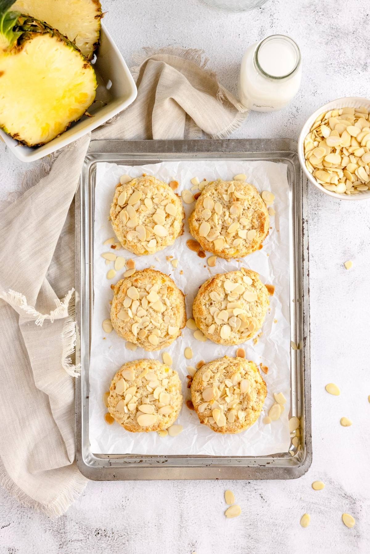 Six almond-topped cookies on a tray with a bowl of sliced almonds, pineapple, and a bottle of milk nearby—perfect inspiration if you're searching for the best fruit scone recipe or want tips for making scones like pineapple scones.
