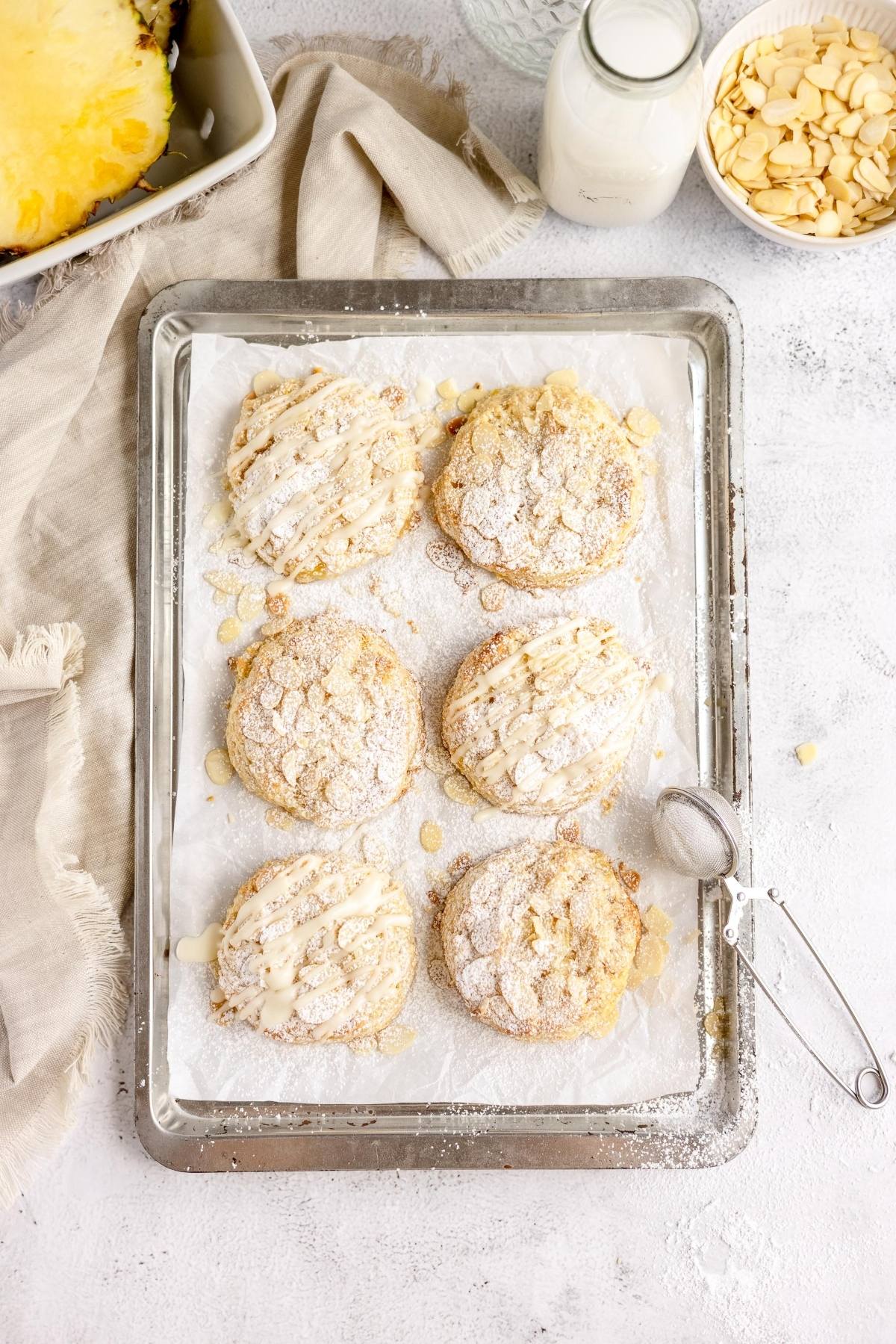 Six almond-topped pastries dusted with powdered sugar on a baking tray, beside milk and sliced almonds—perfect for anyone seeking the best fruit scone recipe or tips for making scones at home.