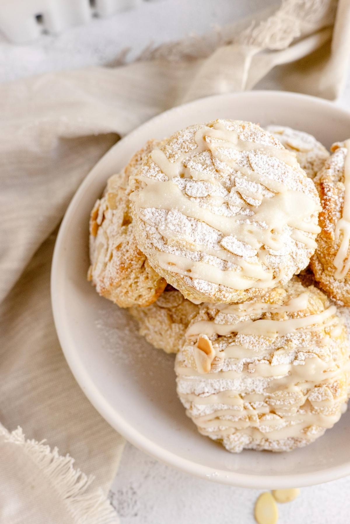 A white bowl of almond cookies topped with powdered sugar and icing on a beige cloth, perfect alongside the best fruit scone recipe or as a sweet twist inspired by tips for making scones.