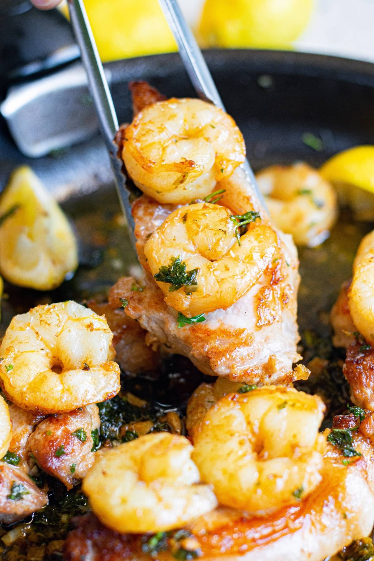 Tongs holding shrimp-topped chicken over a skillet with more shrimp, chicken, and lemon wedges—a delicious surf and turf twist for those who love pork chops and shrimp dishes.