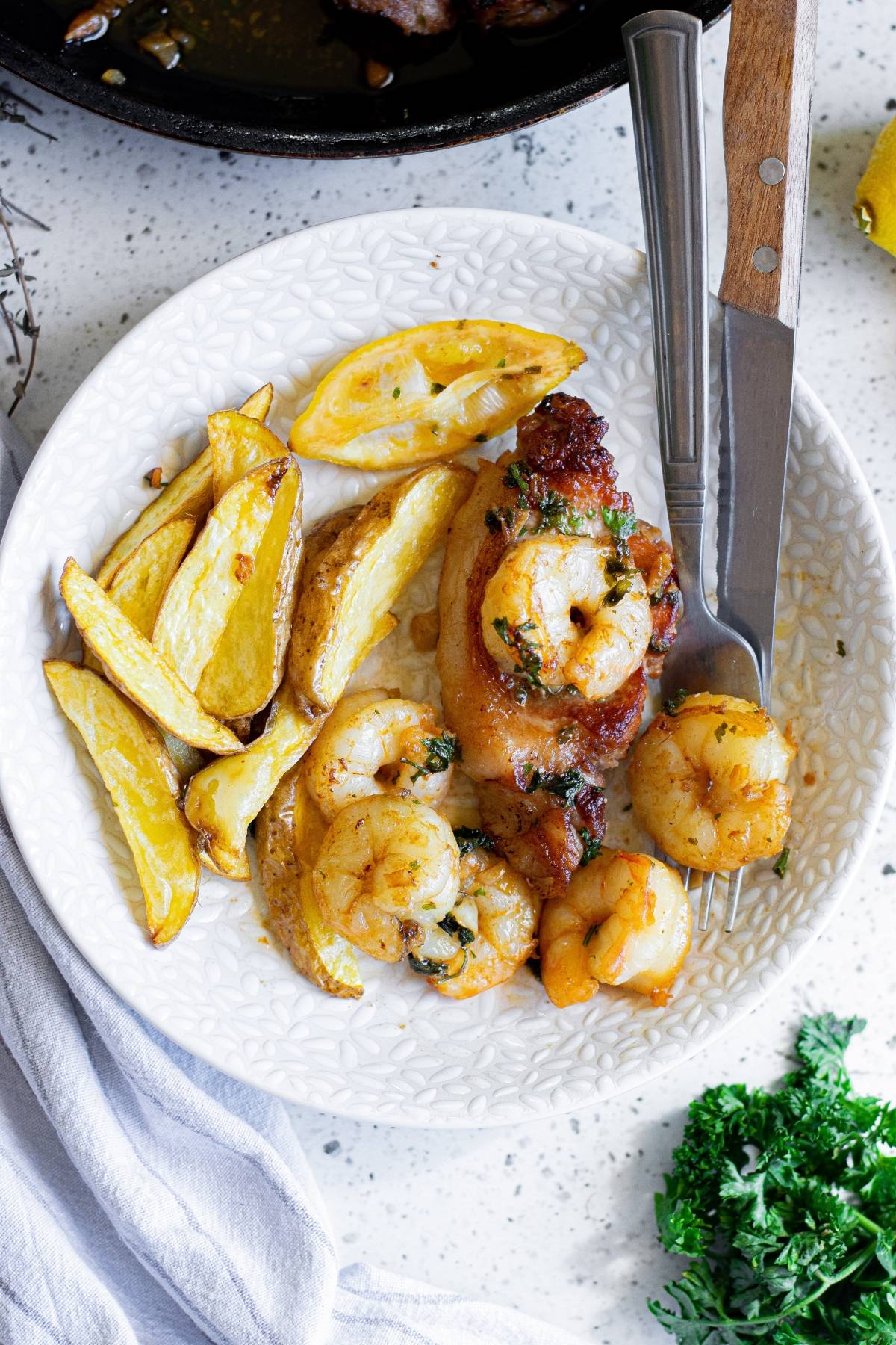 A plate features butter herbed shrimp, chicken, potato wedges, lemon slices, and a fork and knife on the side.