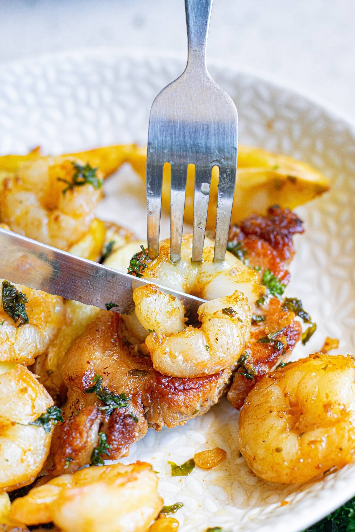 A fork and knife cutting into a seasoned shrimp on a plate with more butter herbed shrimp and herbs.