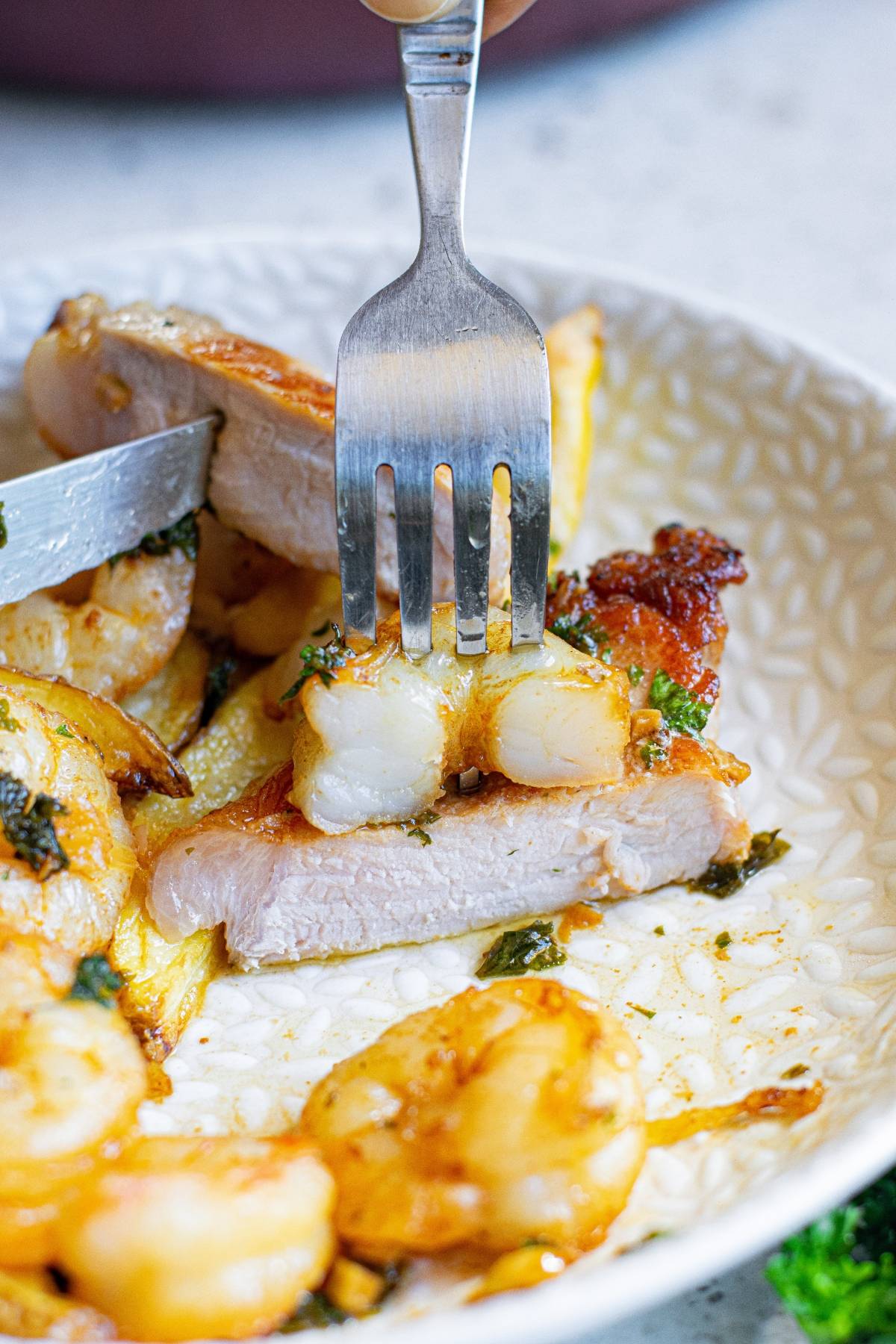 A fork holds a slice of pork chop topped with butter herbed shrimp, served on a plate with more shrimp and fresh herbs—a delicious surf and turf combination.