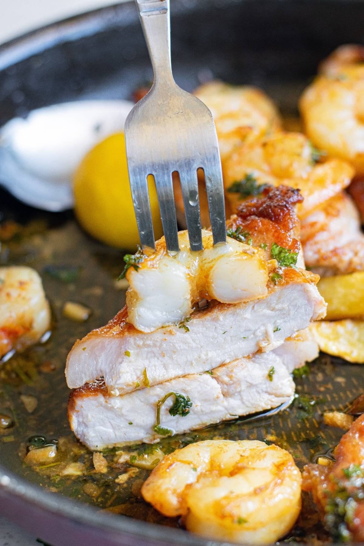 A fork holds a piece of seasoned pork topped with butter herbed shrimp above a pan filled with more shrimp, pork, and a lemon wedge—perfect for trying this easy pork chops recipe.