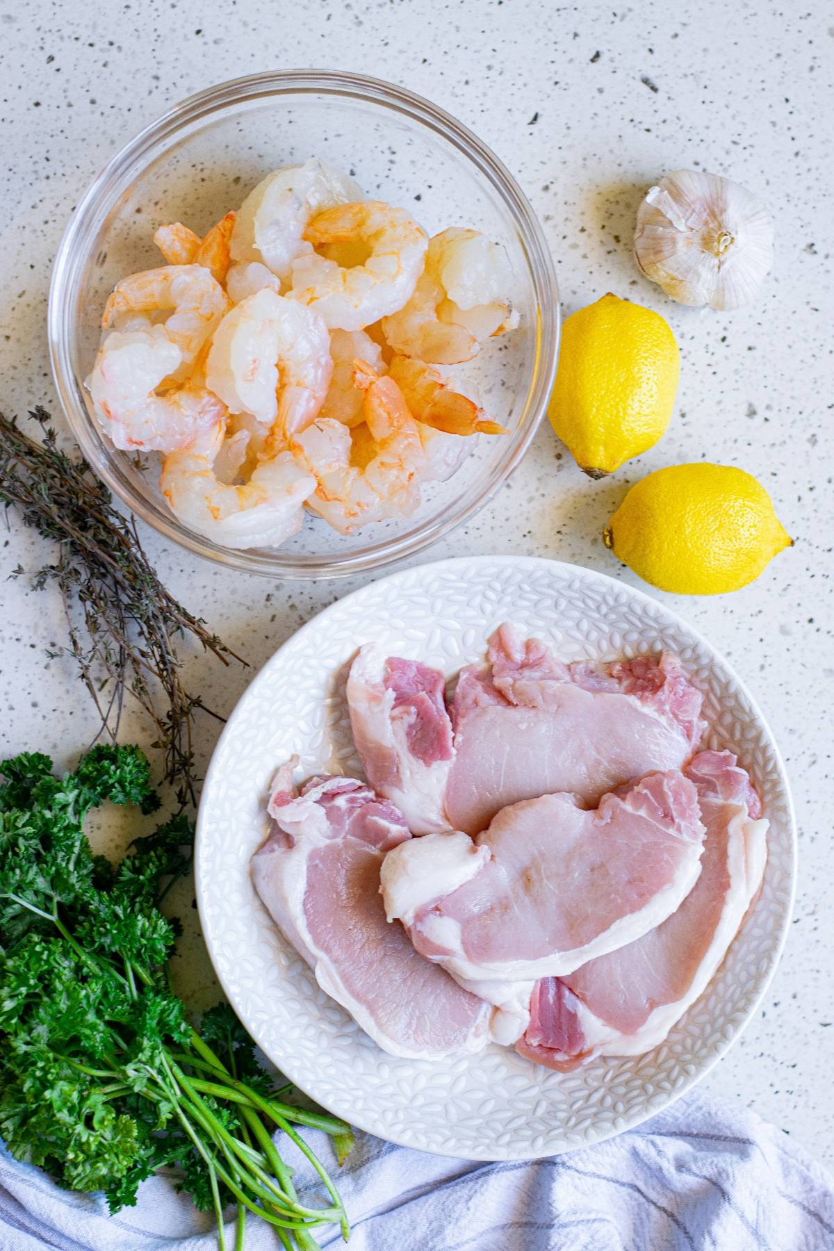 Raw shrimp, pork chops, lemons, garlic, parsley, and thyme arranged on a white countertop—a perfect start for a flavorful pork chops and shrimp surf and turf recipe.