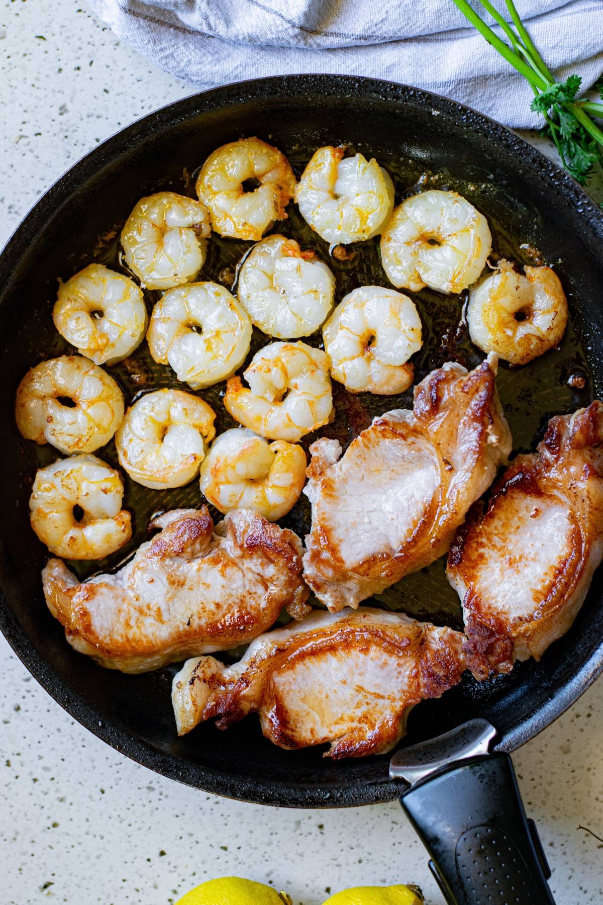 Shrimp and pork chops sizzle in a black skillet on the stovetop, creating a mouthwatering surf and turf meal, with fresh herbs and a towel nearby.