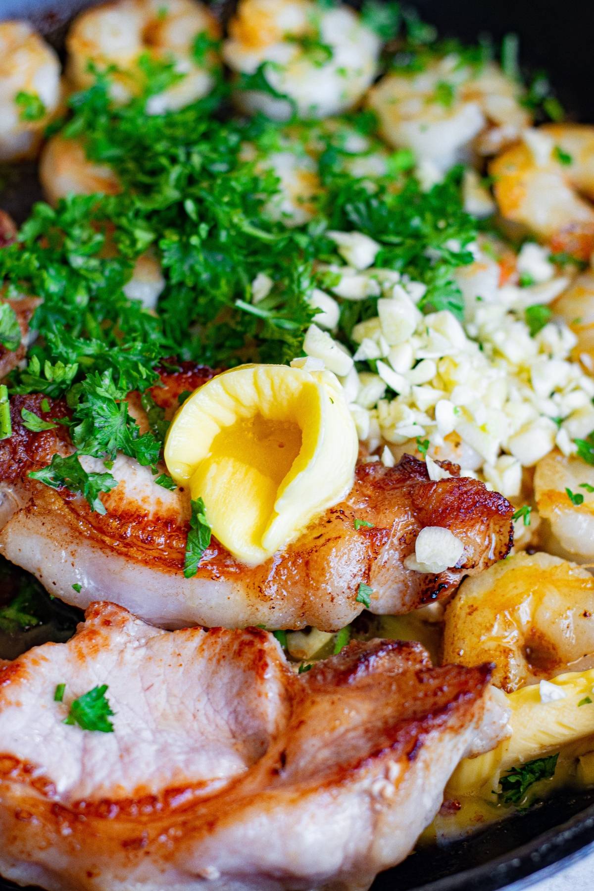 This easy pork chops recipe features pork chops with scallops, chopped garlic, fresh parsley, and a pat of butter in a skillet for a simple surf and turf dinner.