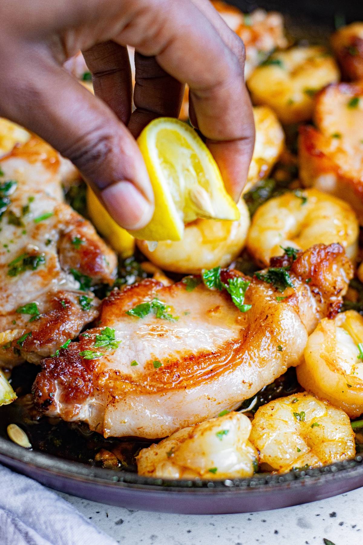 A hand squeezes a lemon wedge over cooked pork chops and shrimp in a skillet with herbs, creating a delicious surf and turf dish.