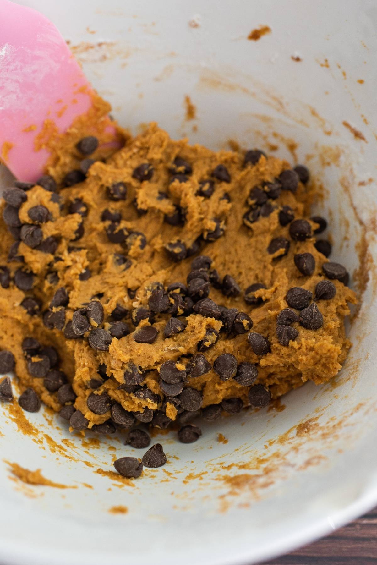 Pumpkin cookie dough with chocolate chips and a pink spatula in a white mixing bowl—proof that pumpkin and chocolate go together for an irresistible treat.