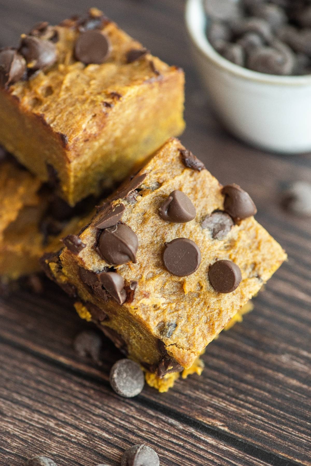 Two pumpkin chocolate chip bars stacked on a wooden surface, with chocolate chips scattered around—proof that pumpkin and chocolate go together perfectly.