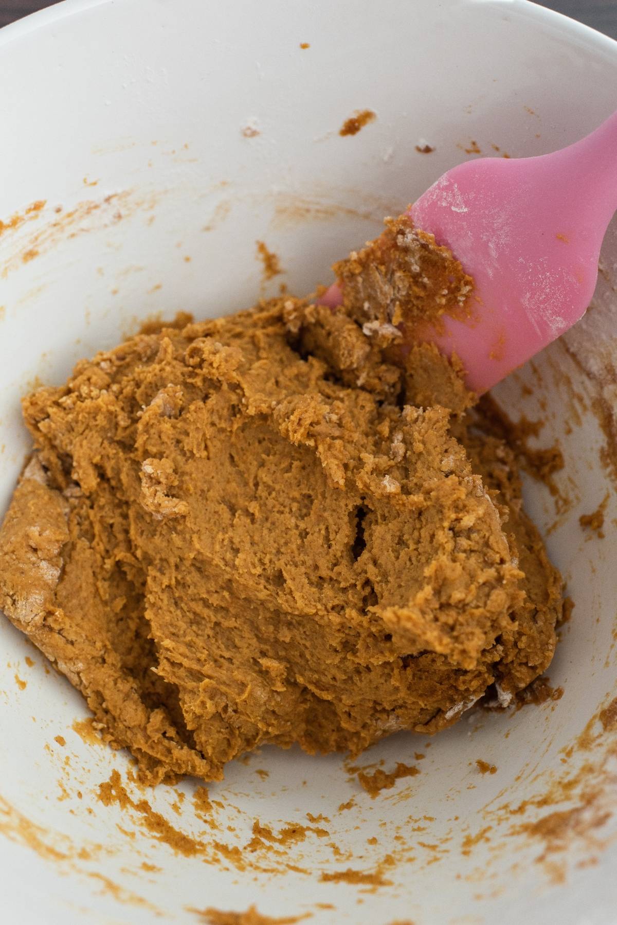 Brown cookie dough in a white bowl with a pink spatula mixing the ingredients, perfect for an easy pumpkin squares recipe or any favorite pumpkin and chocolate recipe.