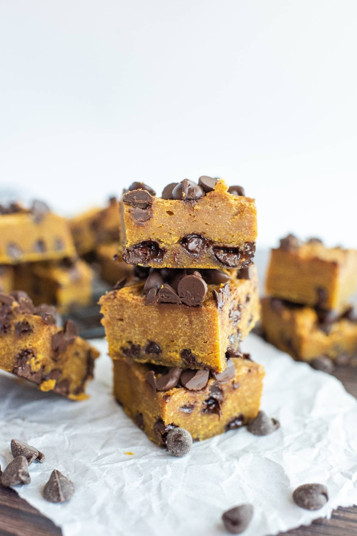 Three stacked pumpkin squares with chocolate chips showcase how pumpkin and chocolate go together, with more bars and scattered chips on parchment paper.