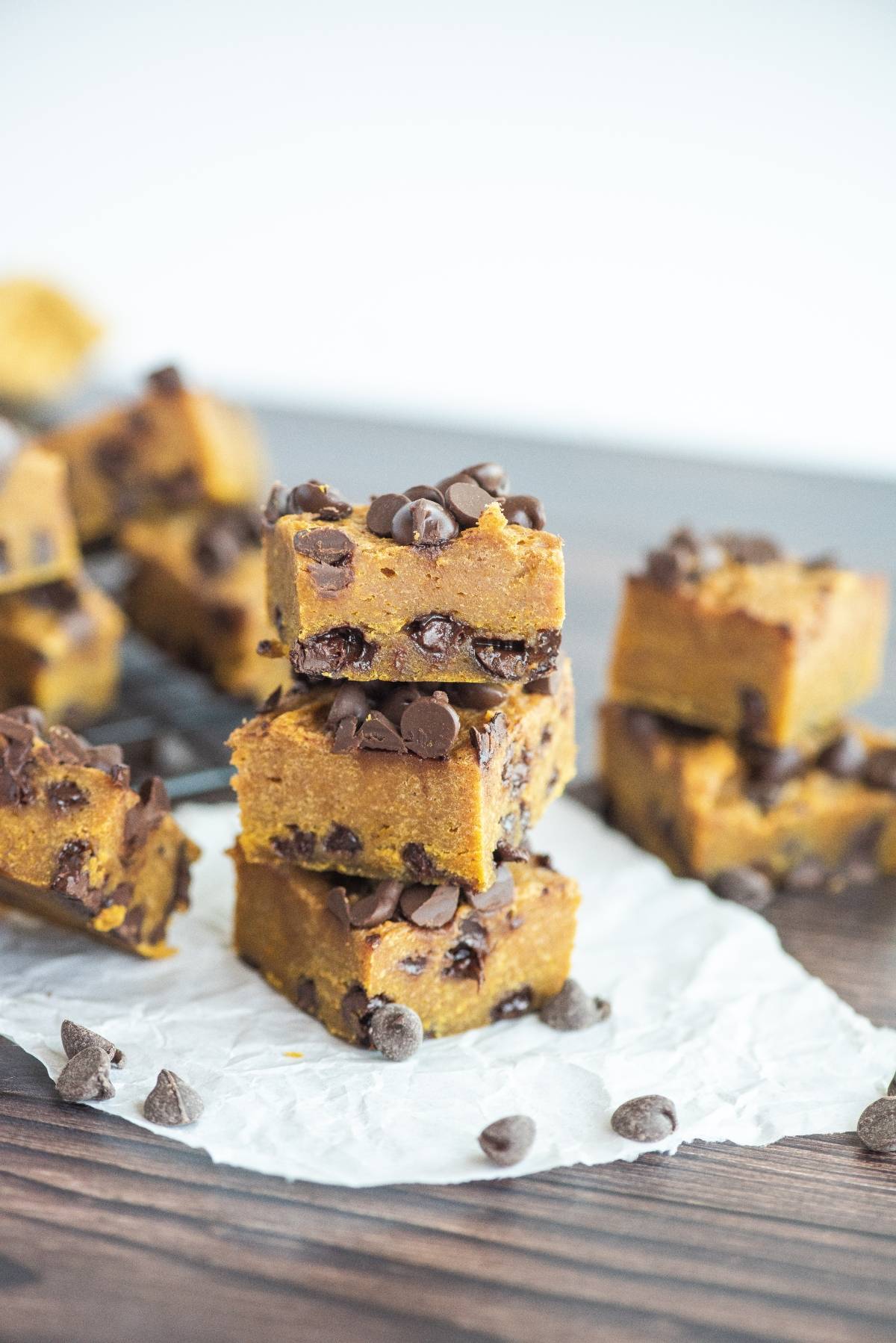 Stacked pumpkin chocolate chip bars on parchment paper, with more bars and chocolate chips scattered around—an irresistible treat for anyone craving an easy pumpkin squares recipe.